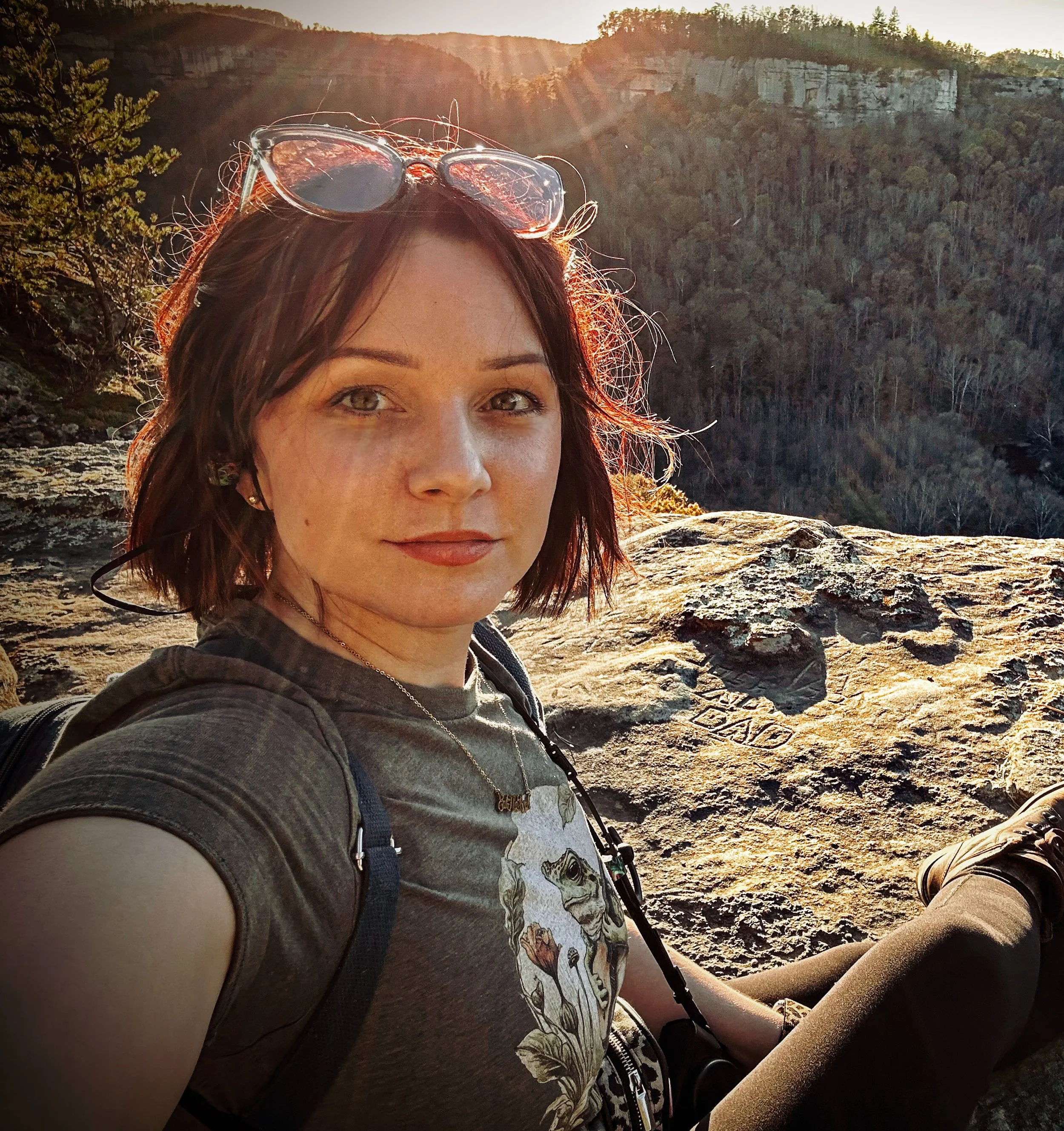 A woman with brown hair and sunglasses on her head takes a selfie in a natural outdoor setting with rocky terrain and forested hills in the background during sunset.