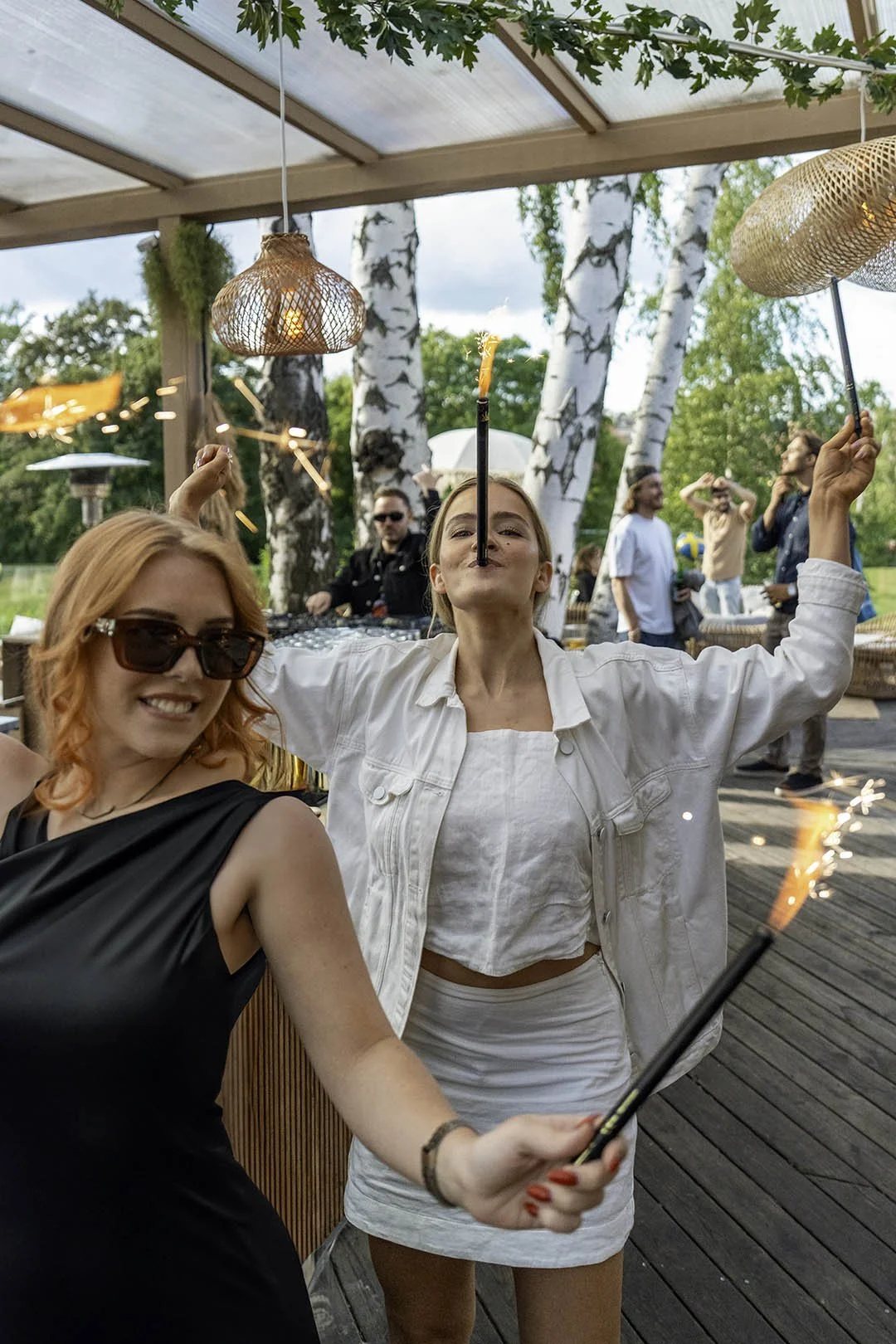 Två kvinnor som leker med tändstickor som avfyrar fyrverkeriliknande små bränder under en fest utomhus omgiven av träd.