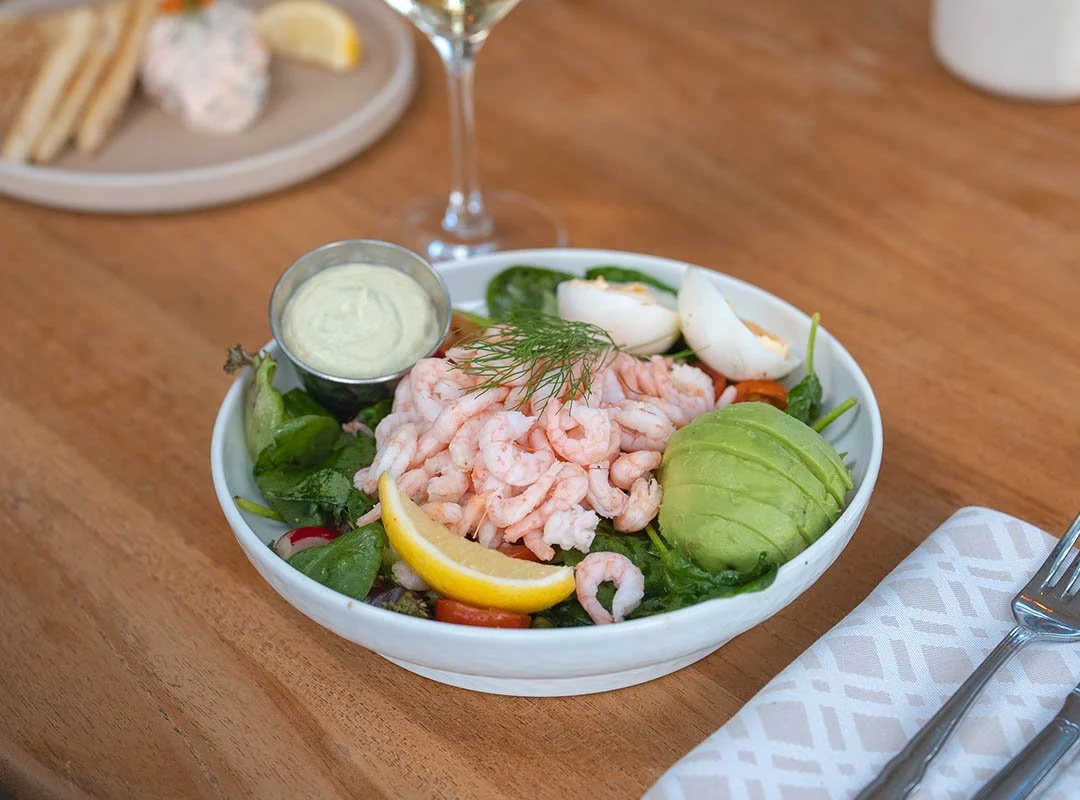 En skål med räkor, kokt ägg, avokado, citron, små bladgrönsaker och en liten bägare med sås, på ett träbord.