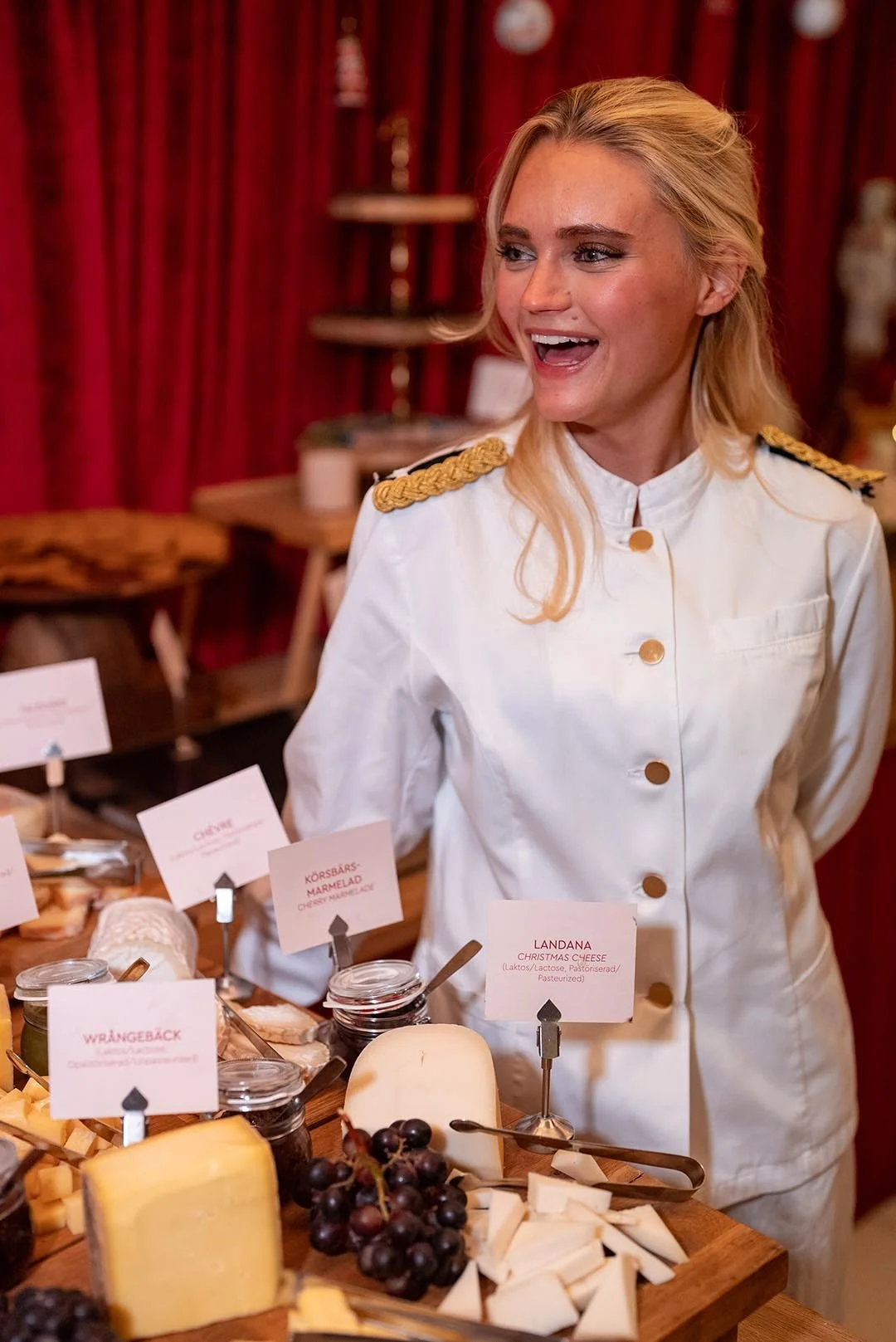 En kvinna i en vit uniform med guldknappar och axelklaffar, står framför en ostbuffé i ett rustikt inrett rum med röda gardiner. Hon ler och tittar åt sidan.