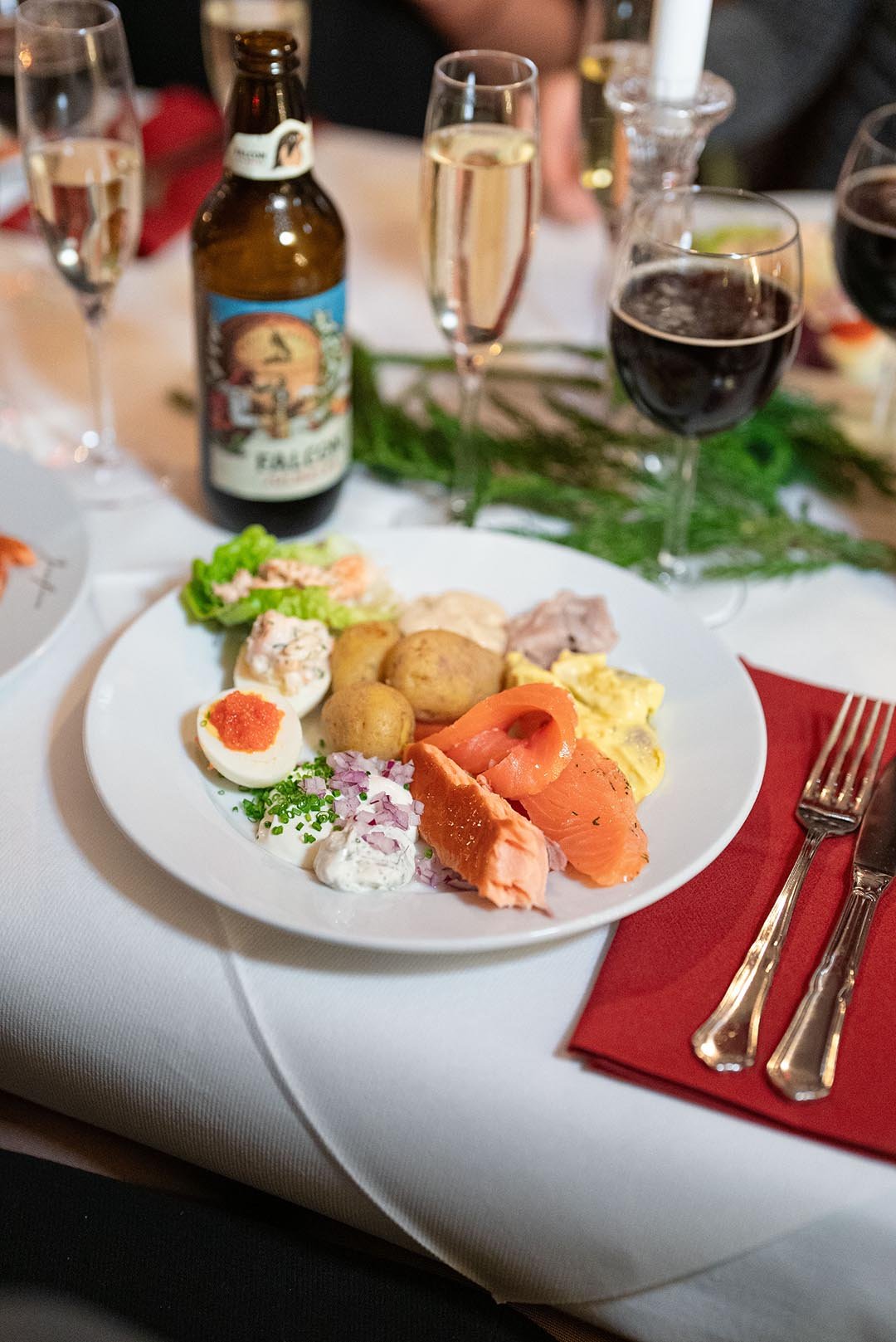 Traditionell julbord med olika sorters sill, lax, köttbullar, ägg, och potatis, drycker som öl, champagne och rödvin på ett festligt bord.