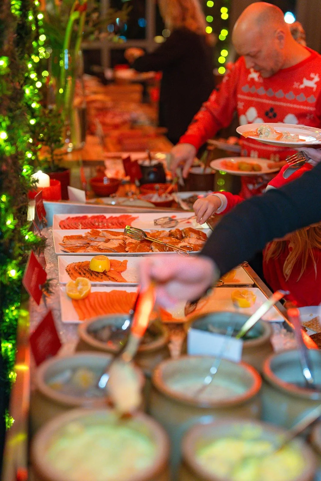 Bild av ett julbord med olika sorters mat, inklusive lax och skaldjur, samt människor som serverar och plockar av mat. Dekorationer och ljus skapar en julig atmosfär.