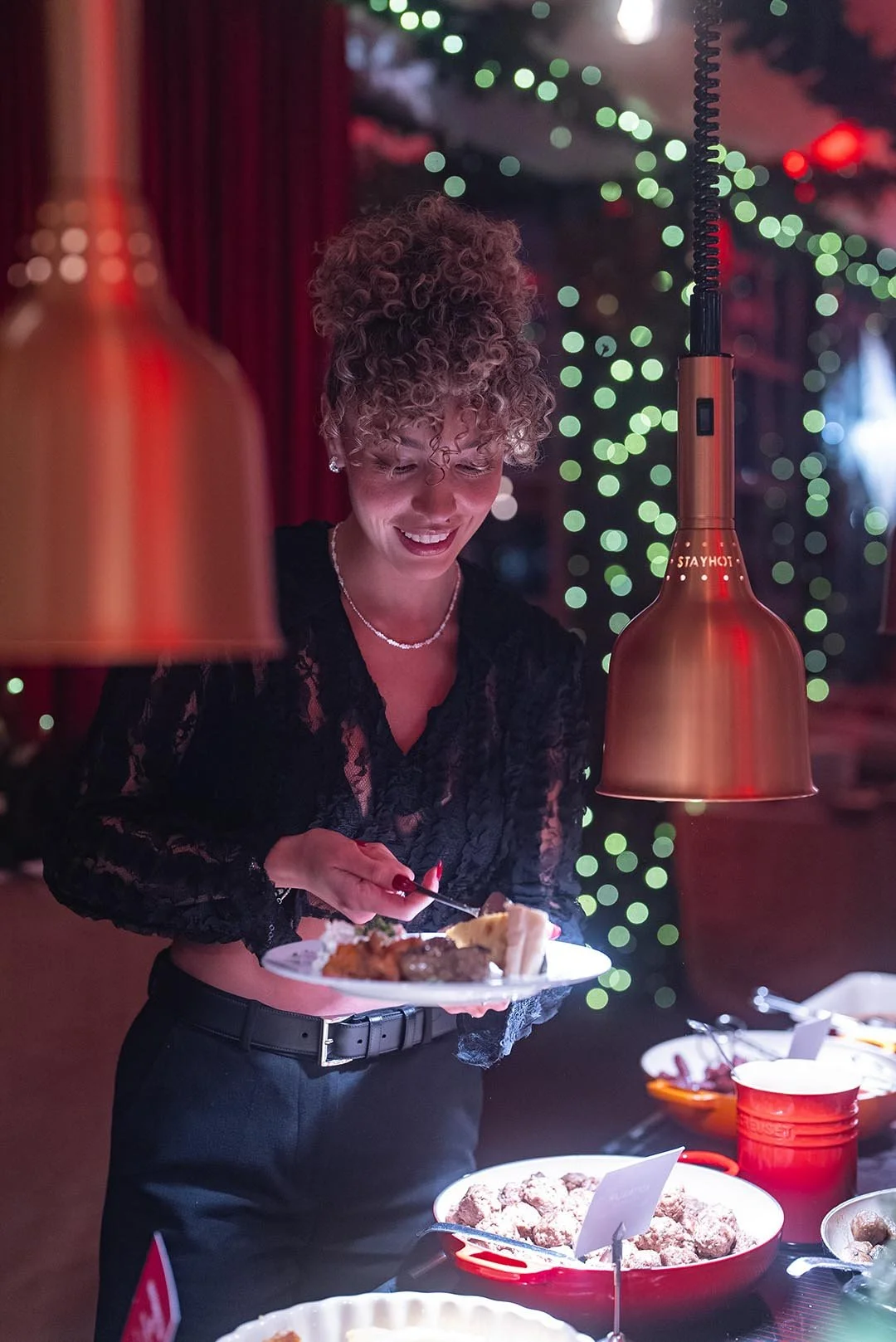 En kvinna i svart klänning serverar mat vid ett julbord, med julgransbelysning i bakgrunden.