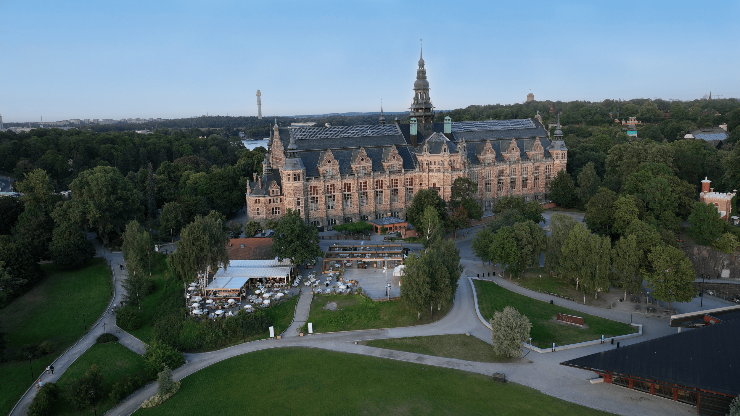 En stor, historisk byggnad som liknar ett slott, omgiven av gröna träd och gröna parker, med en utomhusrestaurang och en liten väg i förgrunden.