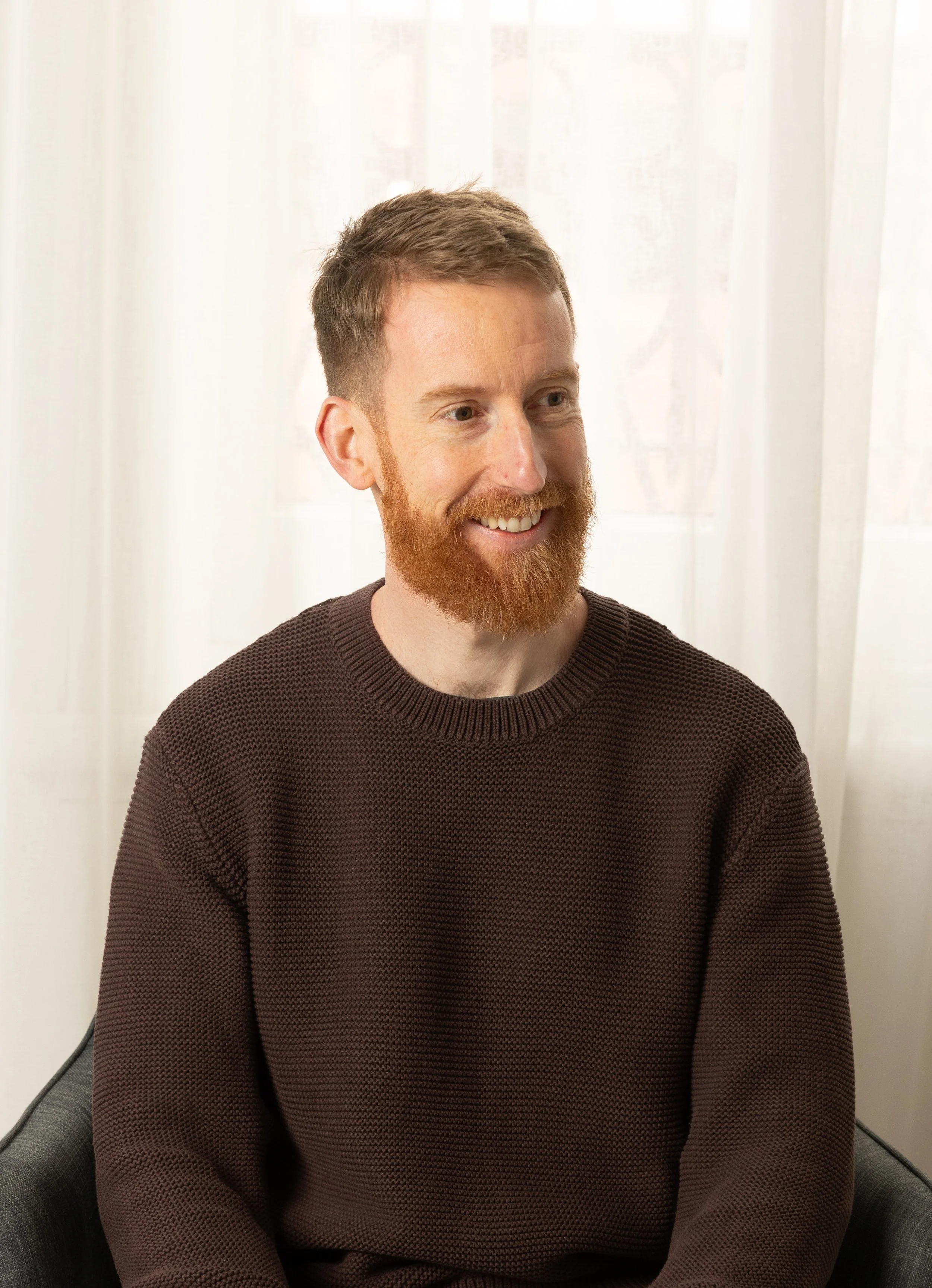 A smiling man with red hair and a beard, wearing a dark brown sweater, sitting on a gray chair in front of a white curtain.