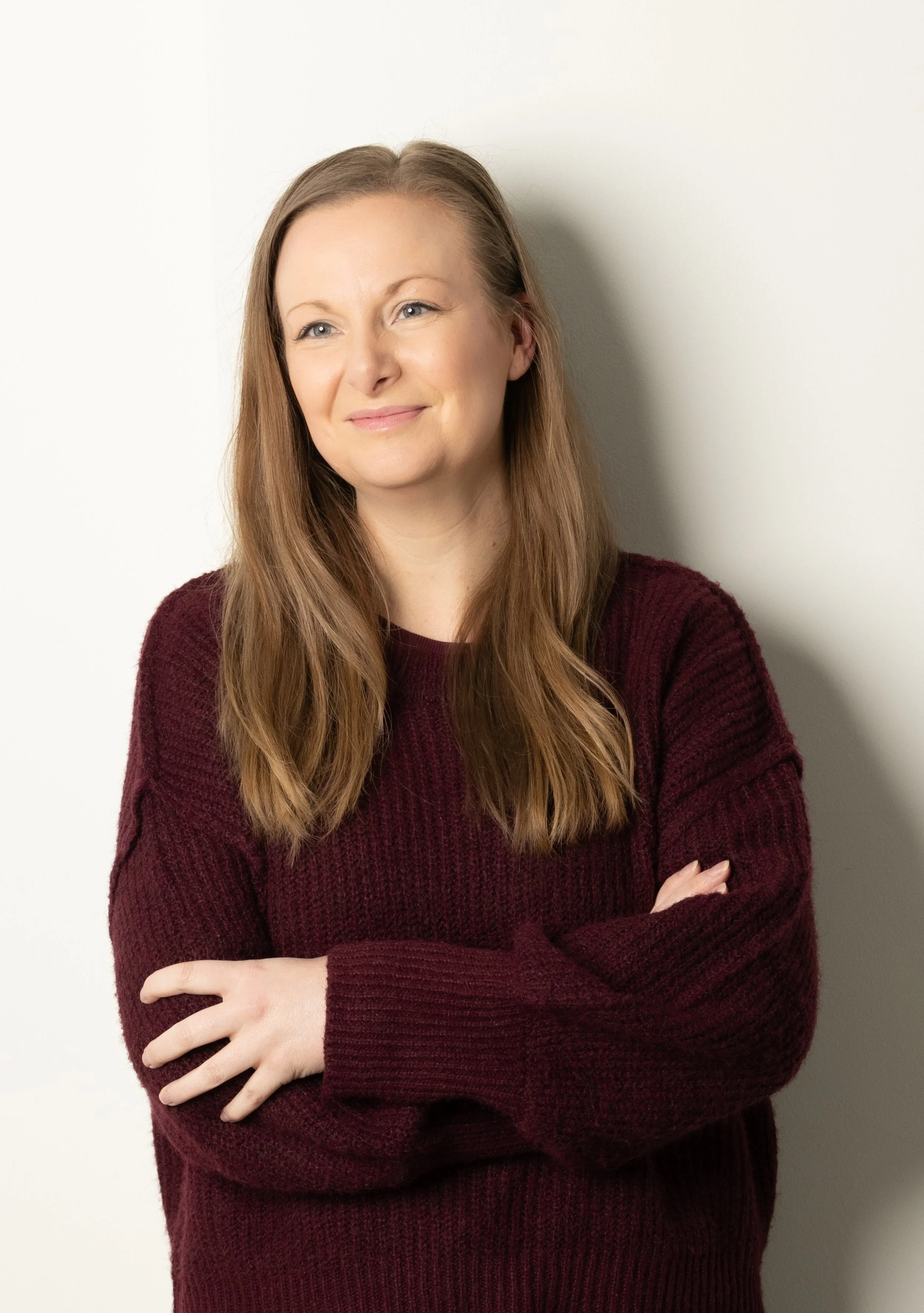 A woman with long light brown hair wears a burgundy sweater and stands against a plain light-colored wall, crossing her arms and smiling softly.