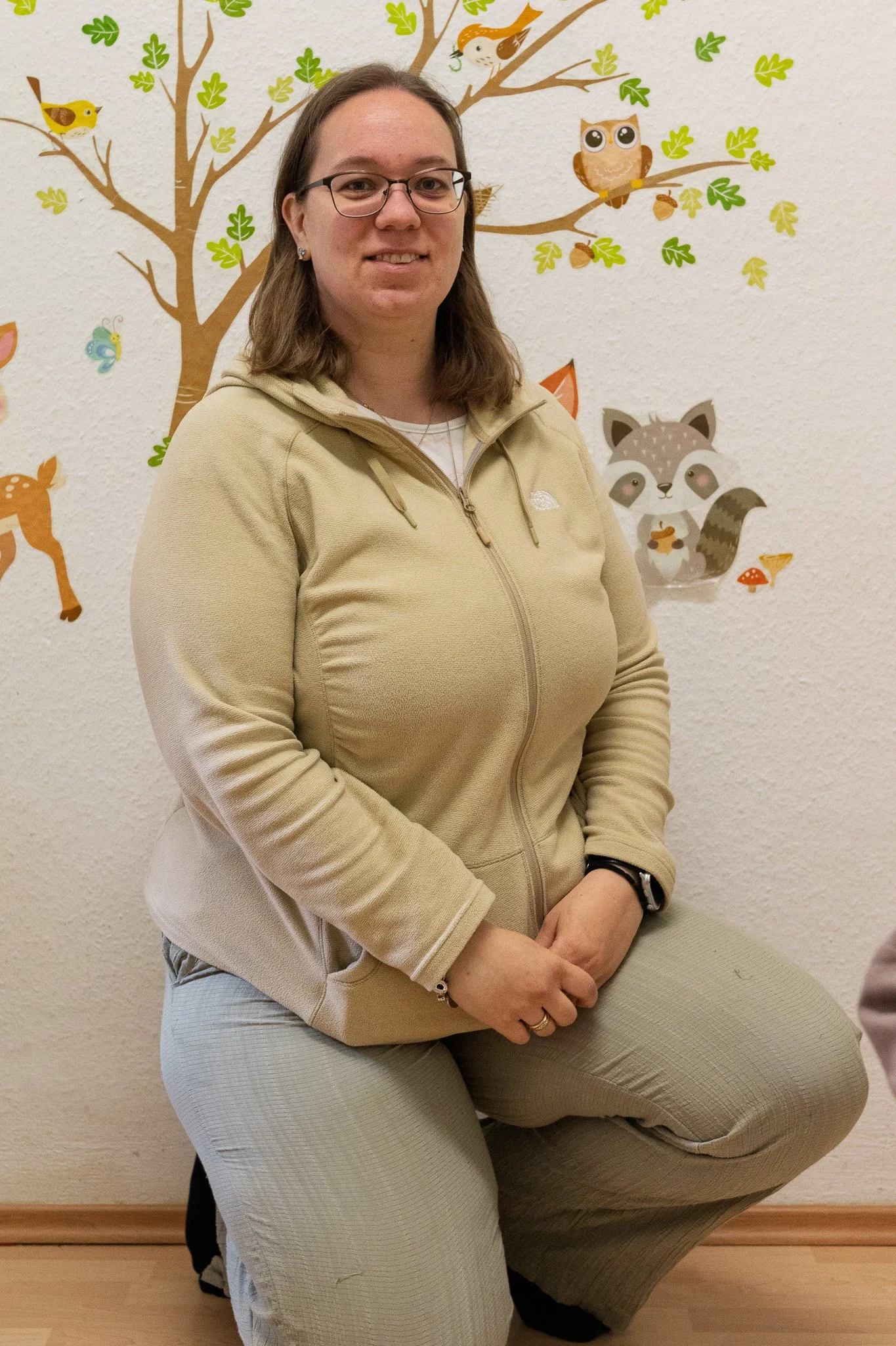 A woman wearing glasses, a beige North Face zip-up jacket, and khaki pants kneeling in front of a colorful woodland animal mural with trees, owls, a fox, a squirrel, and birds on a white textured wall.