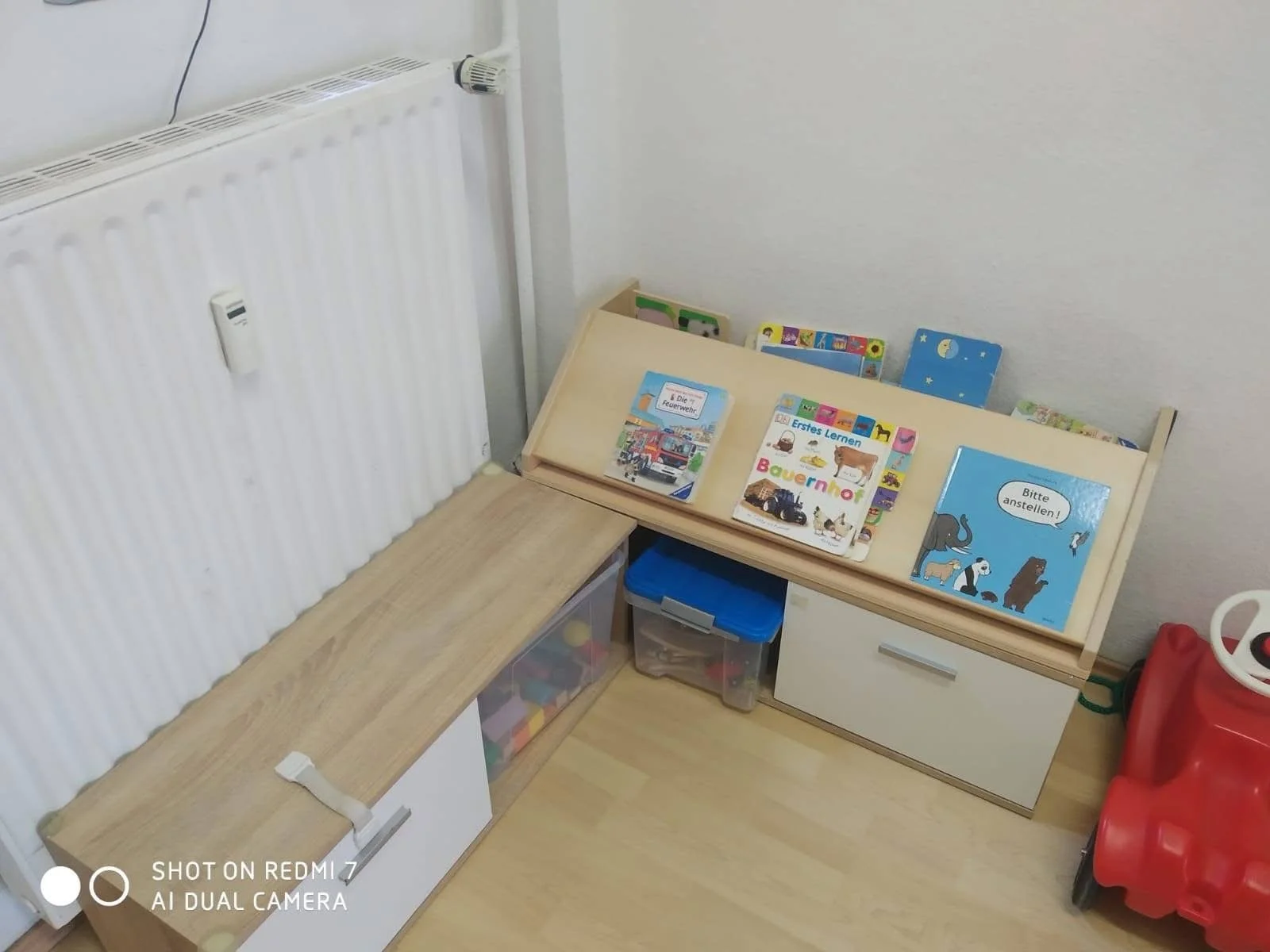 A corner of a room with a wooden book display shelf filled with children's books, a white cabinet below, and a storage bin with colorful toys. There is a radiator on the wall and a red toy ride-on vehicle on the floor.
