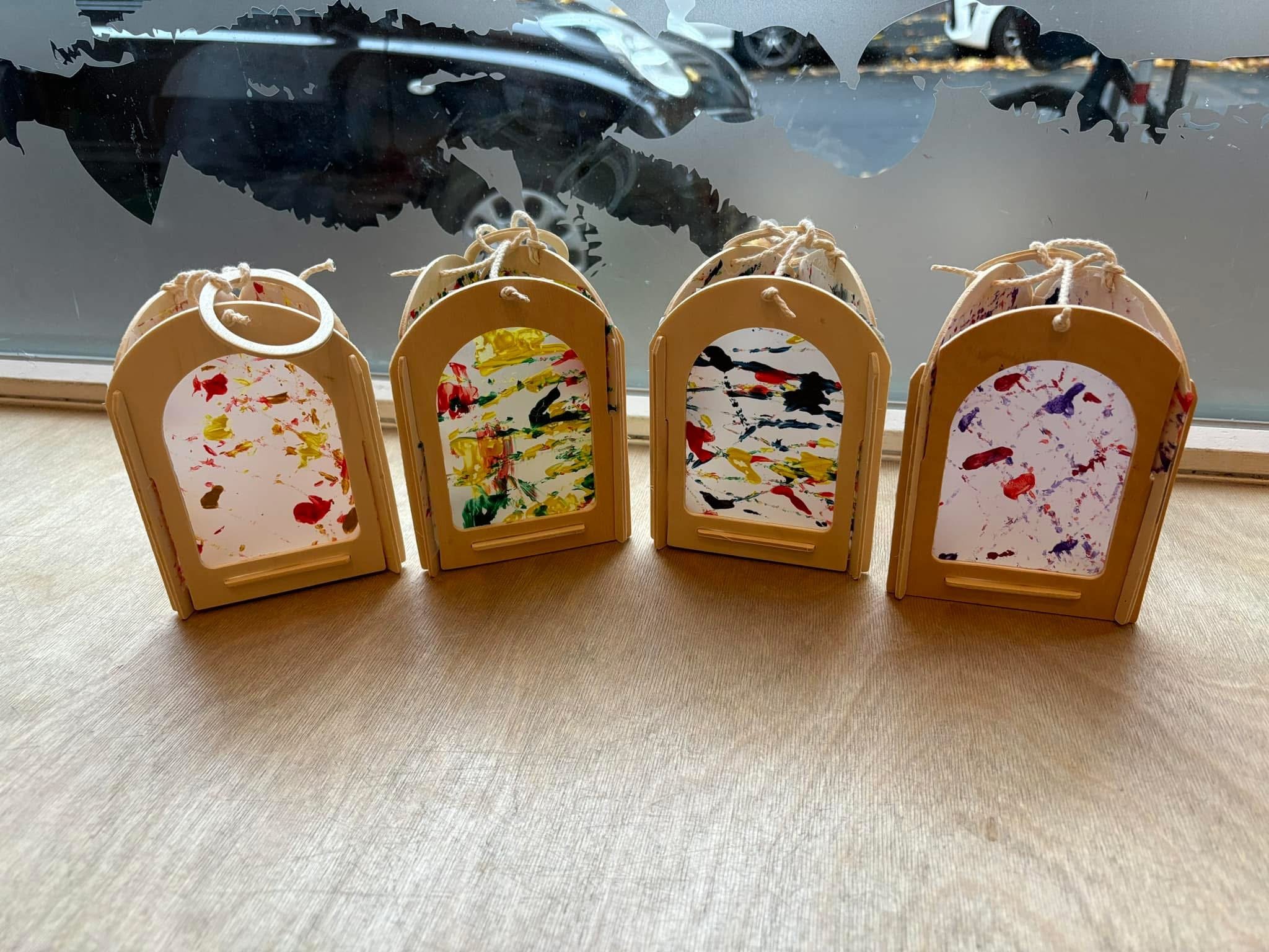 Four small colorful hand-painted hanging art pieces on a window sill with a car and parking lot visible outside.