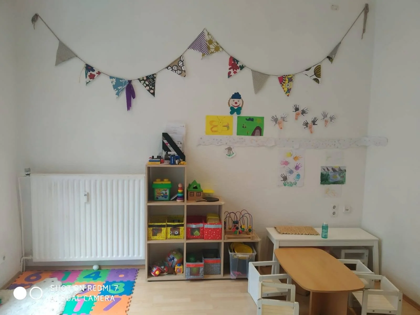Child's playroom with colorful decorations, toys, and furniture. Bunny garland hangs on the wall, and children's drawings are displayed.