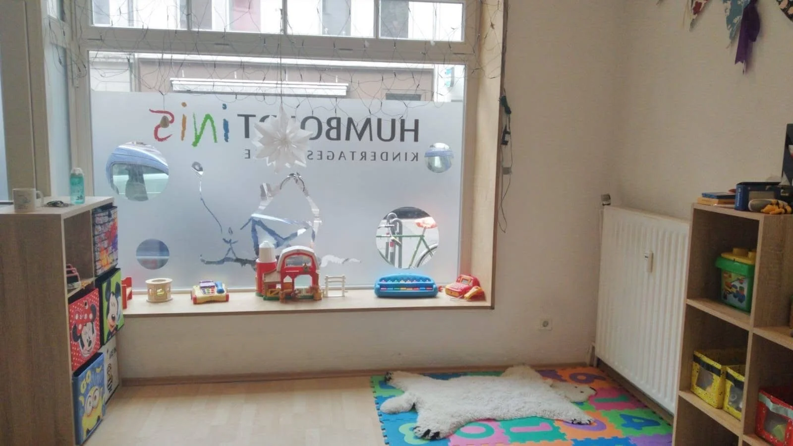 A children's playroom with a window decorated with toys, bookshelves, a colorful foam mat, and a white furry rug.