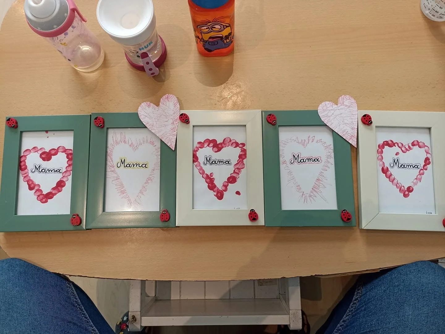 Five picture frames on a wooden table decorated with handwritten and drawn 'Mom' messages inside, surrounded by small ladybug decorations, with pink and white paper hearts attached to the frames.