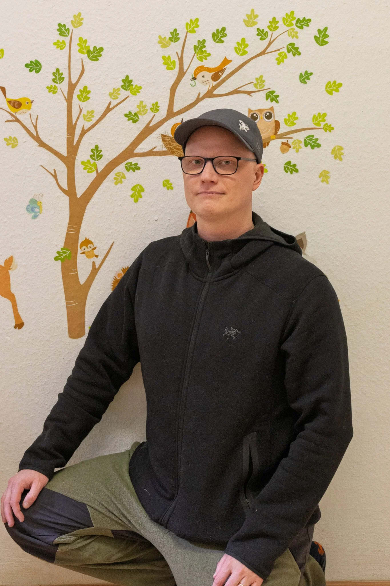 A young man wearing glasses, a black jacket, black cap, and olive-green pants is kneeling with one knee on the ground in front of a wall with a colorful tree and animal wall decal design.