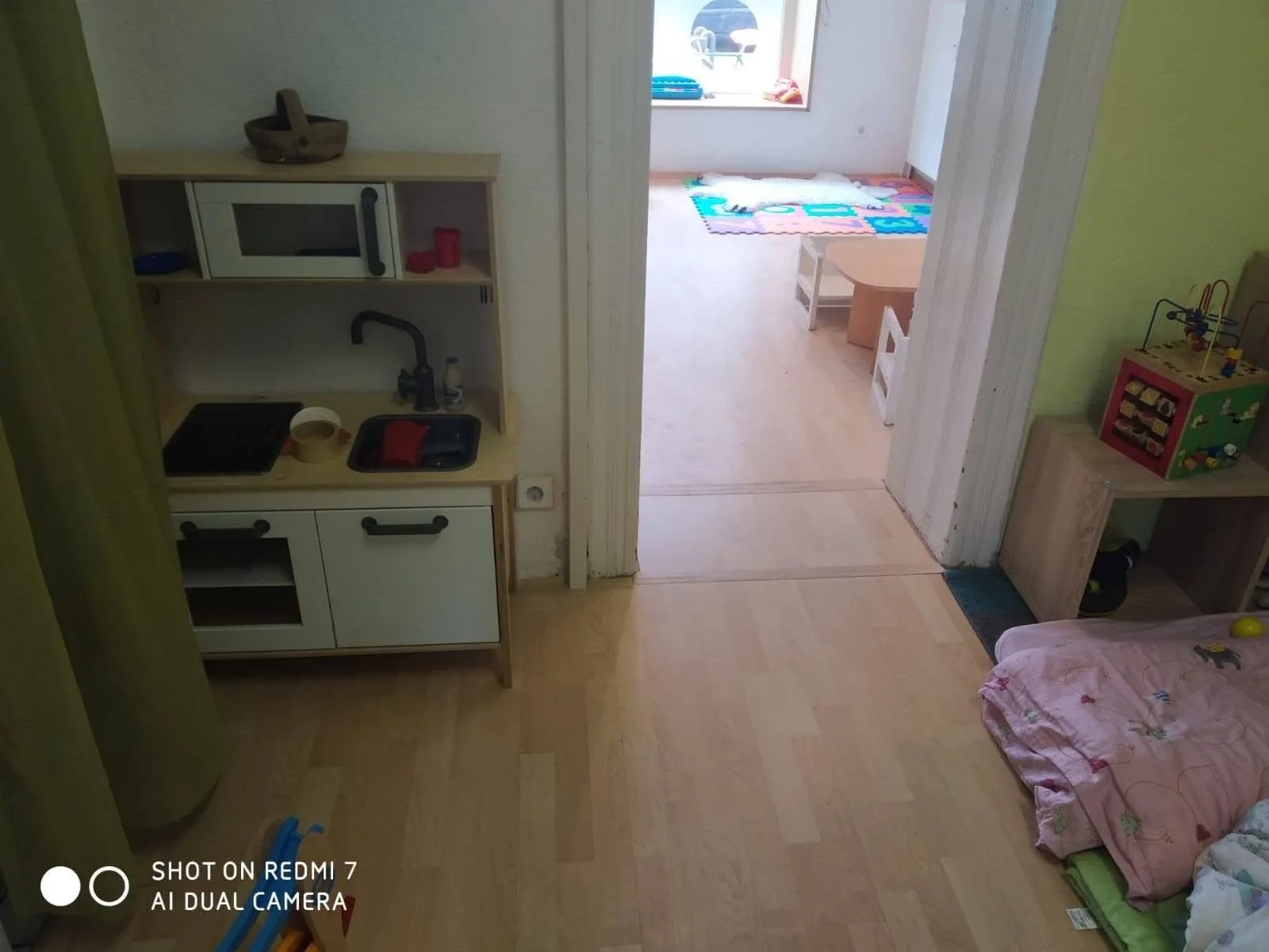 Children's playroom with small beds, toys, a play kitchen, and a colorful rug near the window.