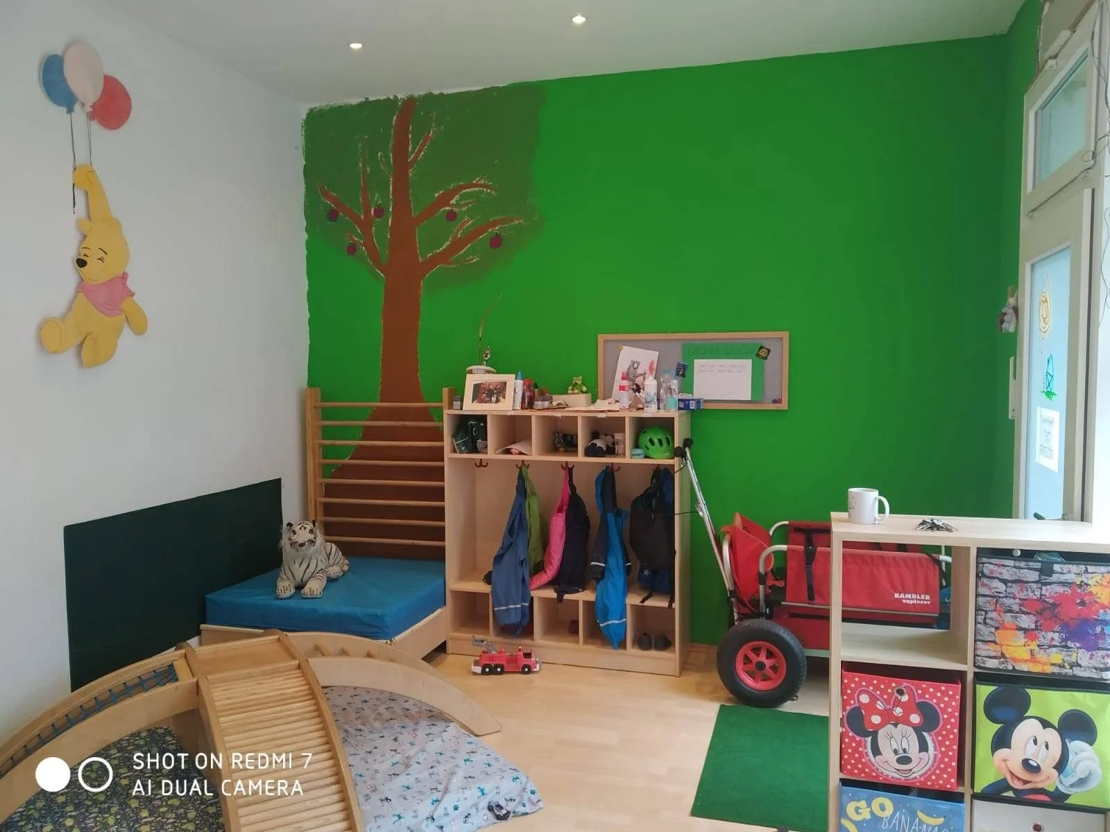 Child's playroom with a mural of a tree on the green wall, a wooden shelf with backpacks, a red wagon, Mickey Mouse themed storage cube, and a bed with a striped plush tiger toy.