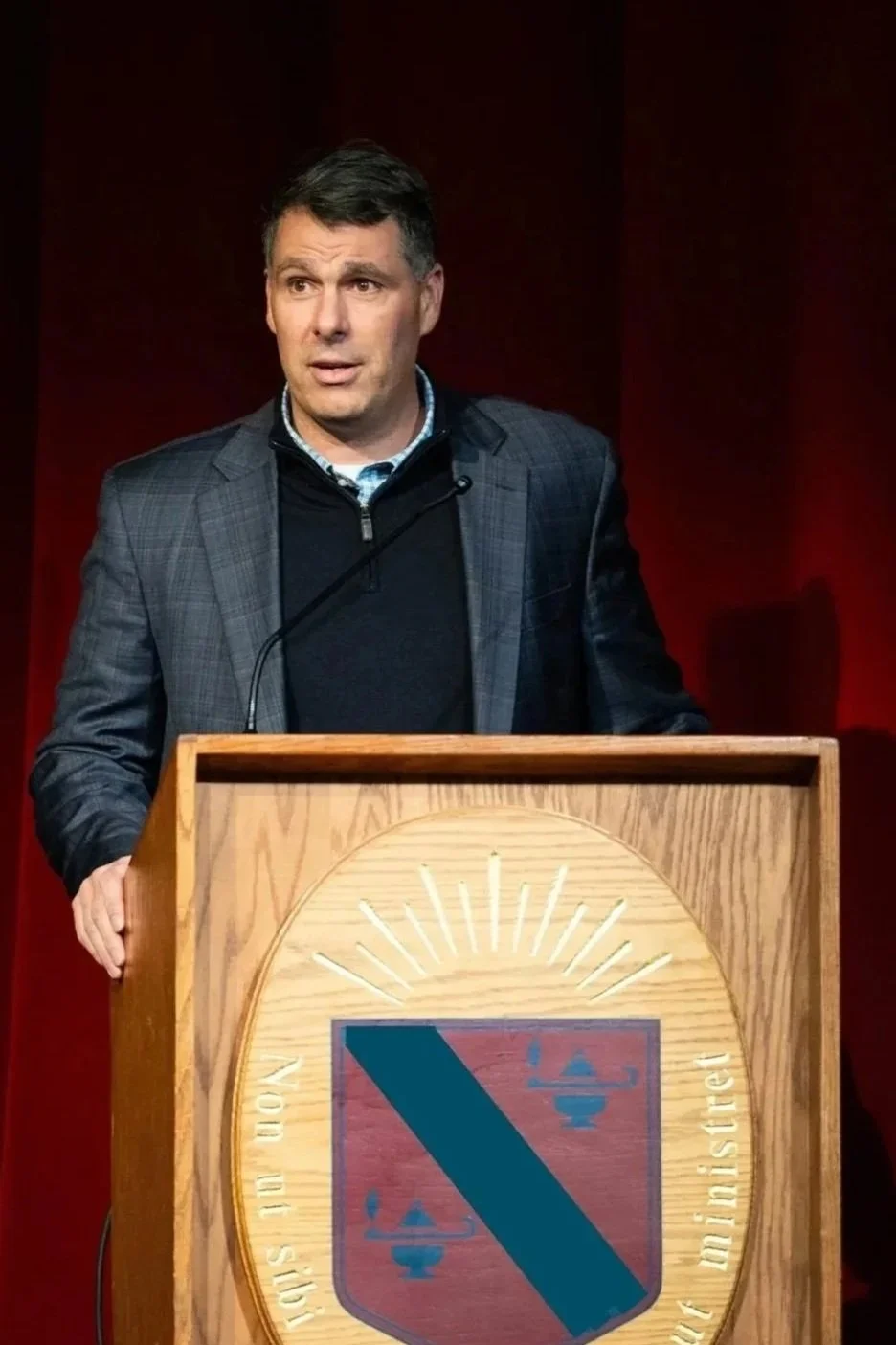 A man in a checked blazer and black sweater giving a speech at a wooden podium with a red and blue crest, against a red background.