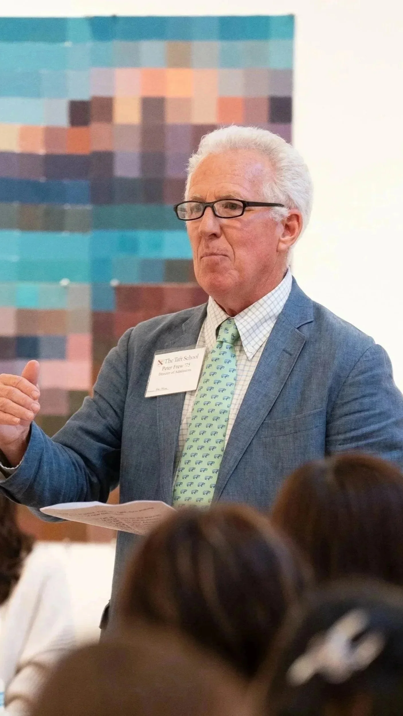 An elderly man with white hair, glasses, dressed in a suit with a patterned tie, is speaking or presenting in front of a group, holding papers. There is a blurred pixelated artwork in the background and an audience in the foreground.