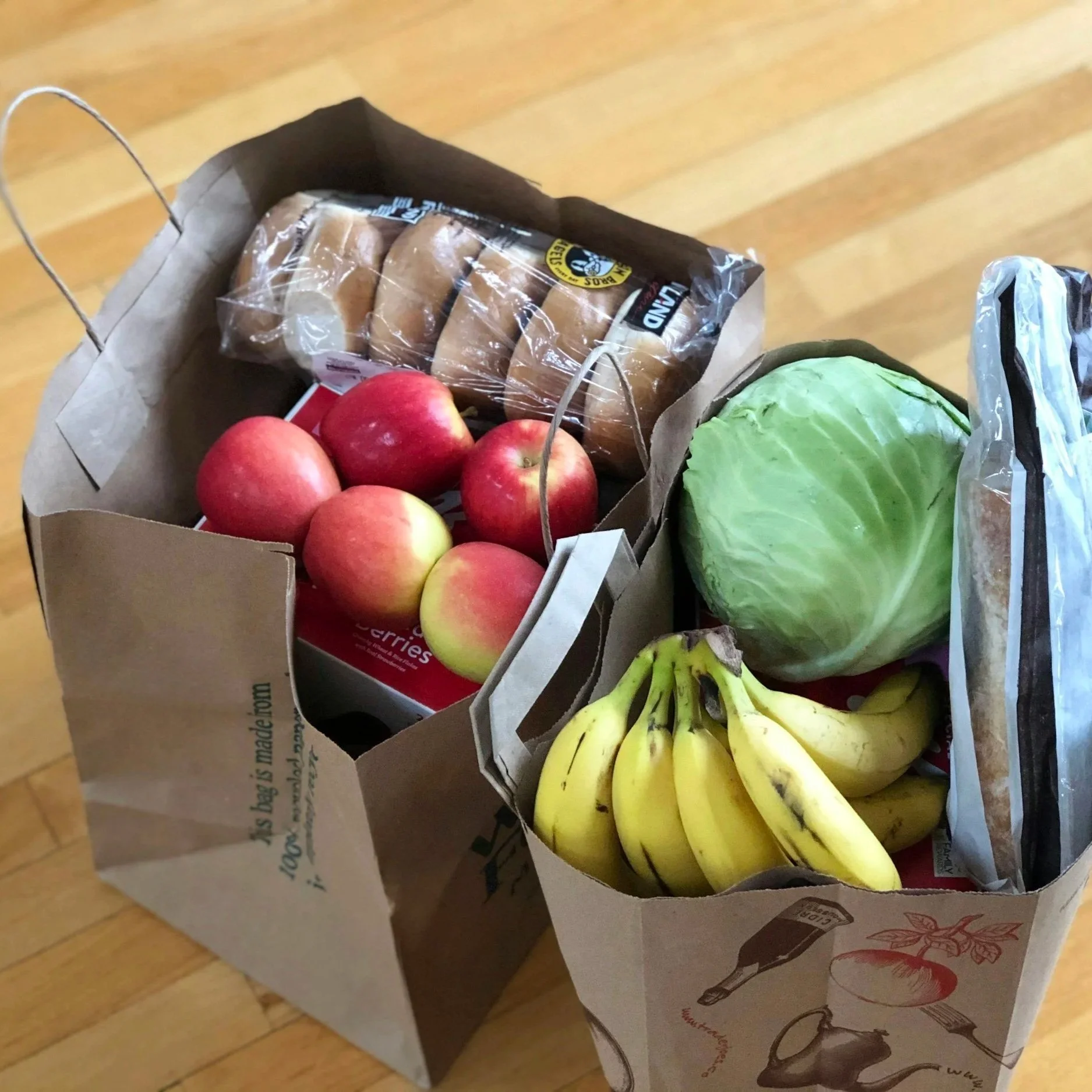 A paper grocery bag containing red apples, a head of green lettuce, a bunch of bananas, a bag of potato buns, and a bag of sliced bread, sitting on a wooden floor.