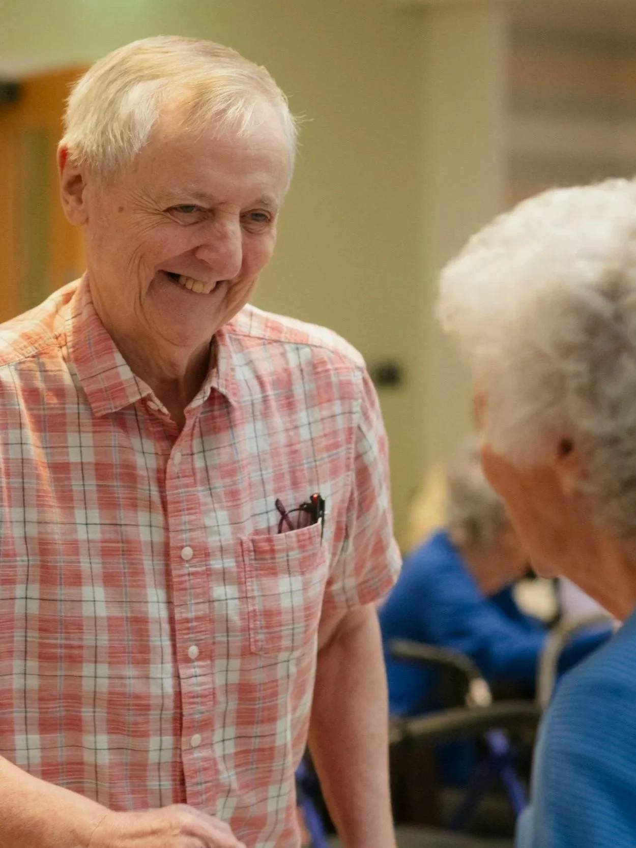 Smiling senior man wearing a pink plaid shirt, talking to a woman with gray hair in a busy indoor setting.