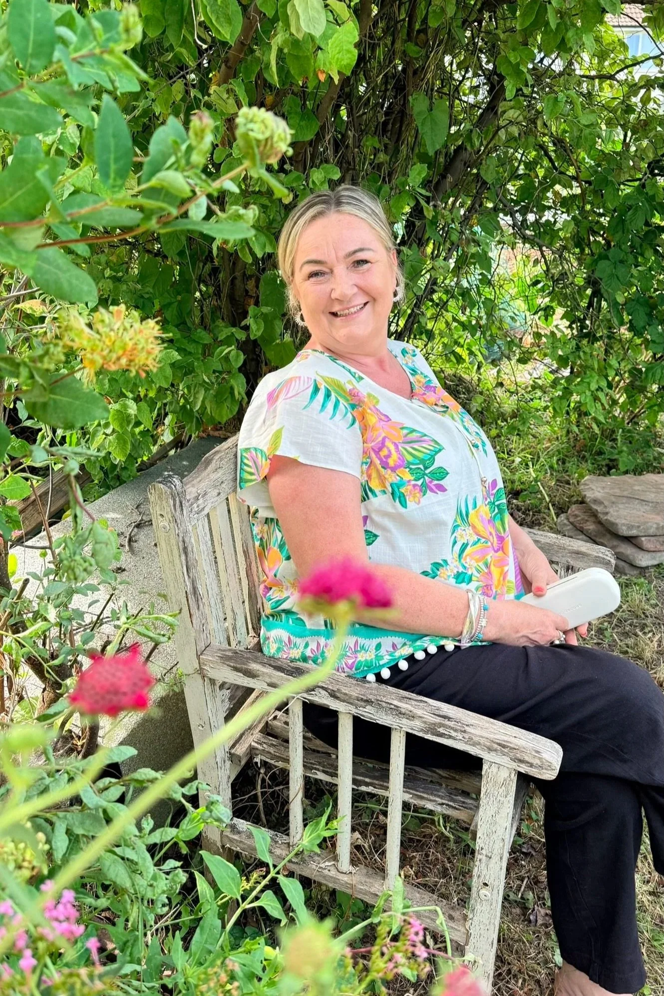 A smiling woman sitting on a weathered wooden bench outdoors surrounded by green foliage and pink flowers, wearing a colorful floral shirt and black pants.