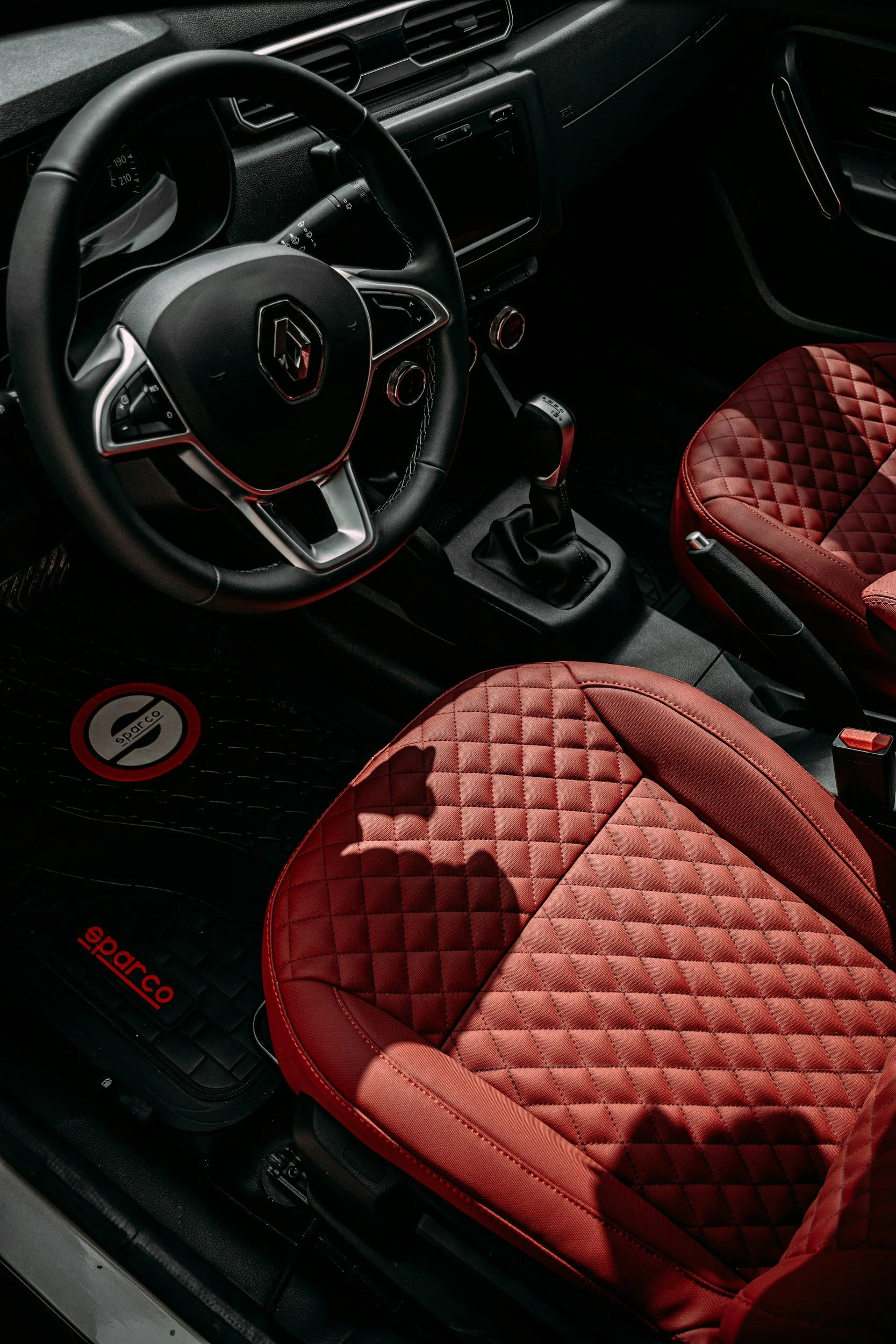 The image shows the interior of a car, featuring a black steering wheel with a logo, a touchscreen dashboard, gear shift, and red quilted seat covers on the driver's and passenger's seats. The car floor has a black mat with a red and white logo.