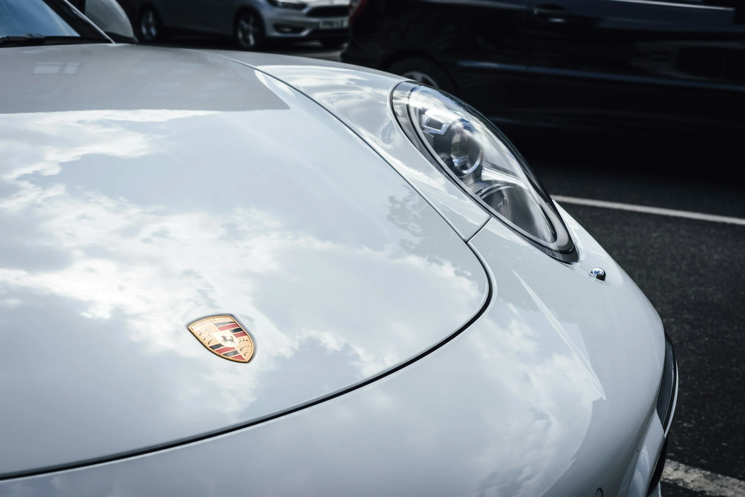 Close-up of the front of a silver Porsche sports car parked in a parking lot, with the Porsche emblem visible on the hood.