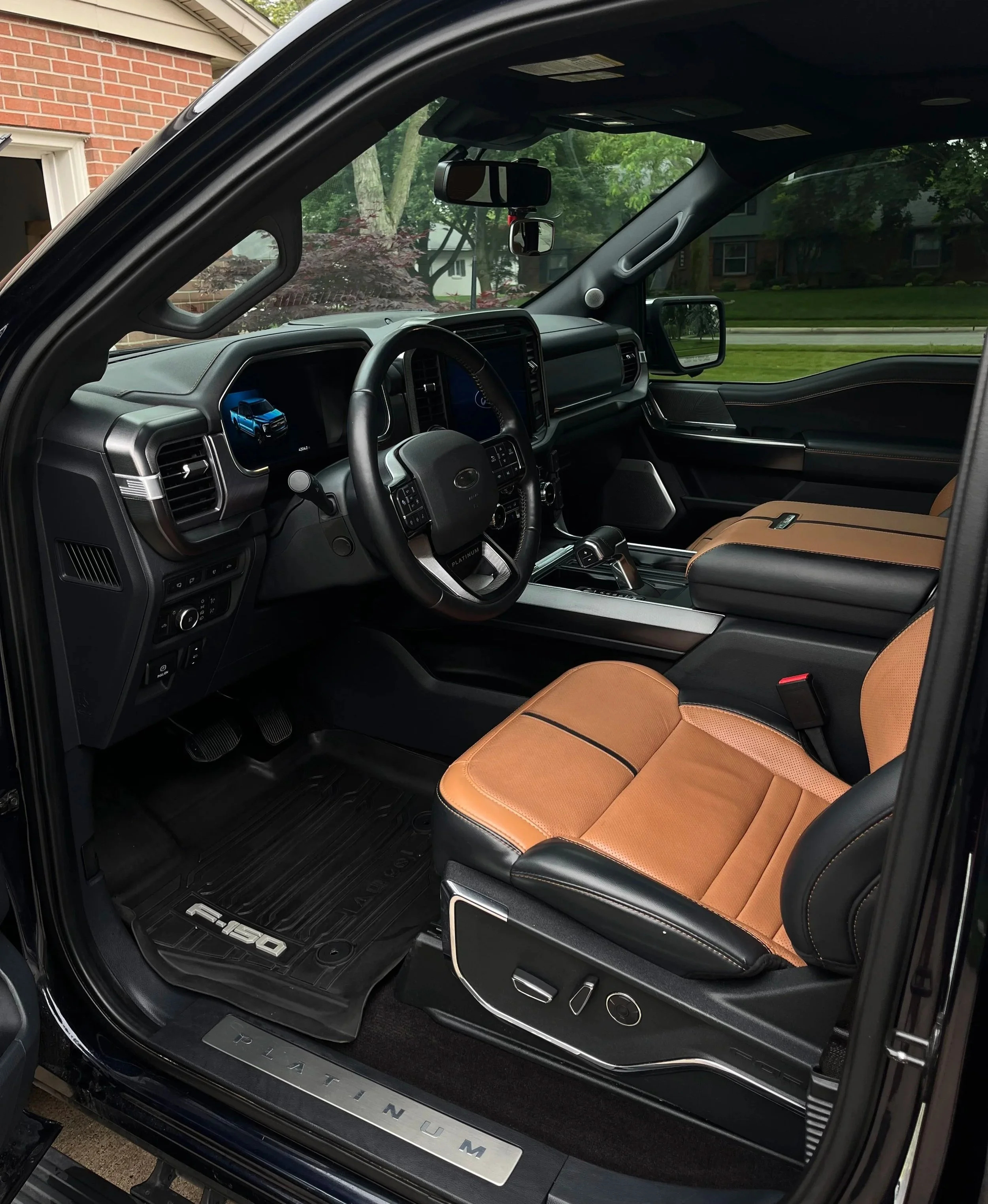Interior of a Ford F-150 pickup truck with tan leather seats, black dashboard, and centered infotainment screen, showing the vehicle's interior and part of a residential area outside.