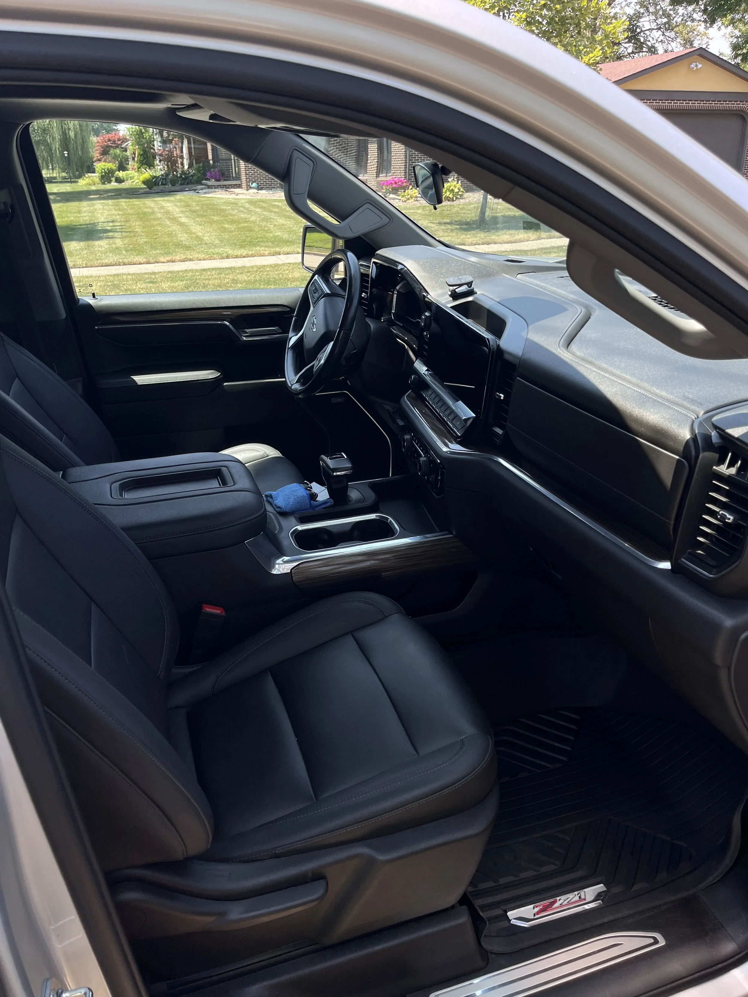 Interior view of a black pickup truck showing front seats, dashboard, steering wheel, center console with gear shift, and dashboard touchscreen, with a residential neighborhood visible through the windows.