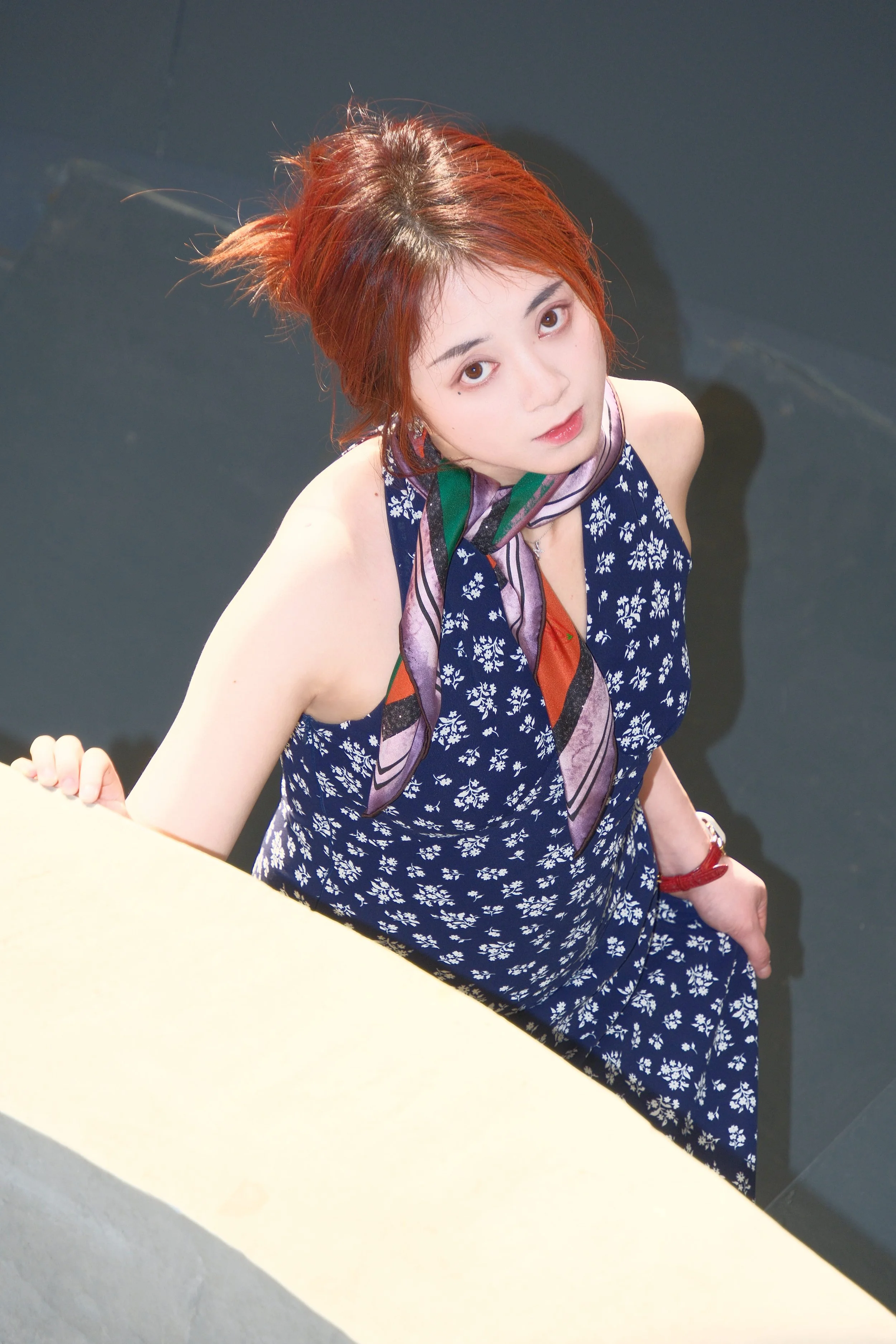 A young woman with short, reddish-brown hair looking up at the camera. She is wearing a navy blue dress with white floral patterns and a multicolored scarf around her neck. The background is dark, possibly a wall.