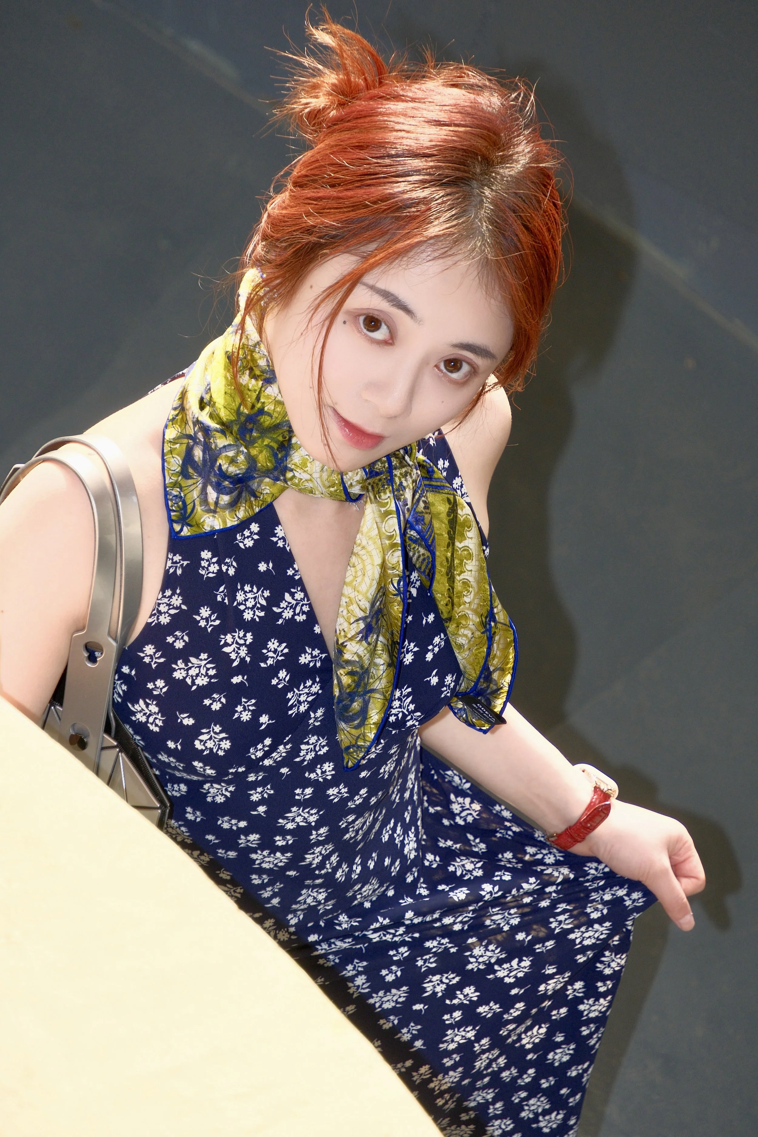 A young woman with reddish hair styled in a messy bun, wearing a navy blue dress with a white floral pattern, and a yellow scarf with blue patterns around her neck. She has brown eyes, a small mole near her eye, and is sitting at a table. She is pointing with her right hand, wearing a red bracelet. The background is dark and blurred.