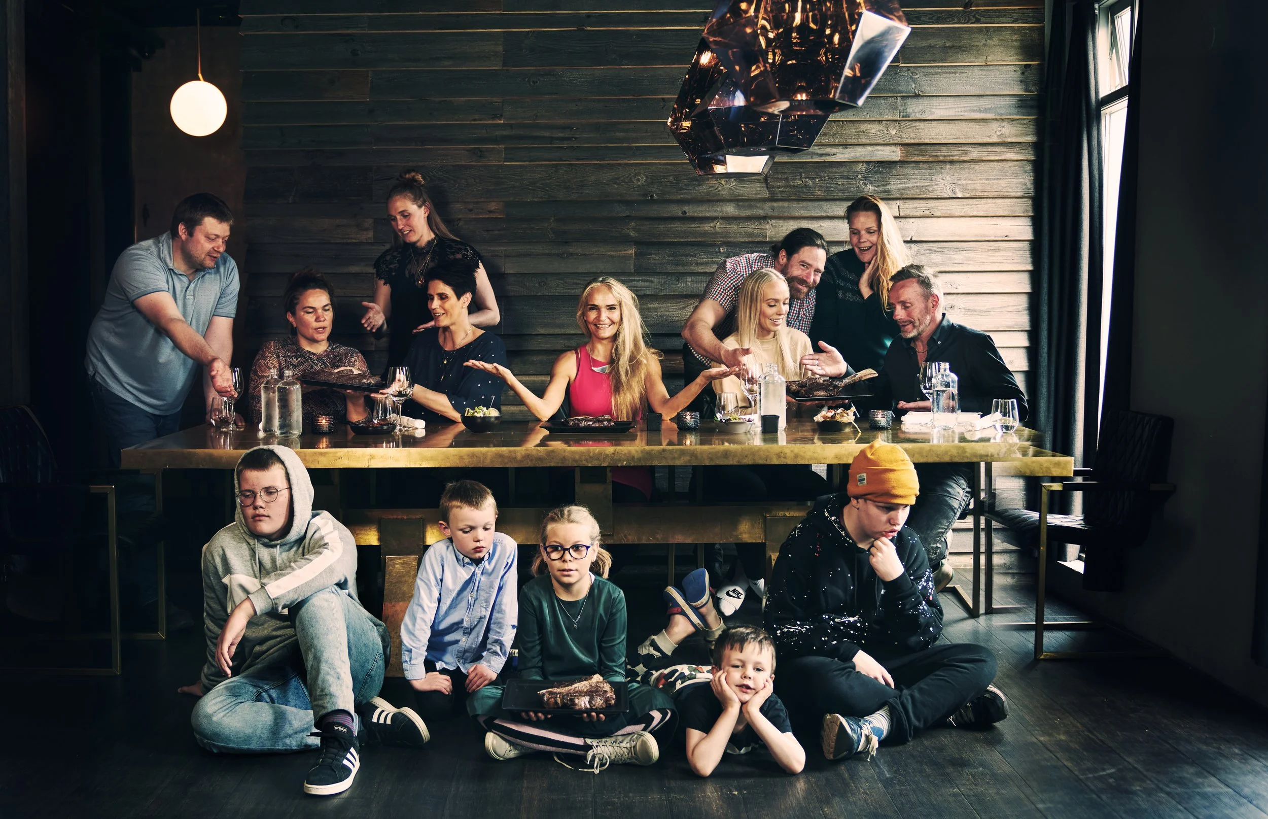 A group of children sitting on the floor and people sitting and standing at a long table in a dining area with wooden walls, some eating, some talking, and some looking down, with various drinks and food on the table.