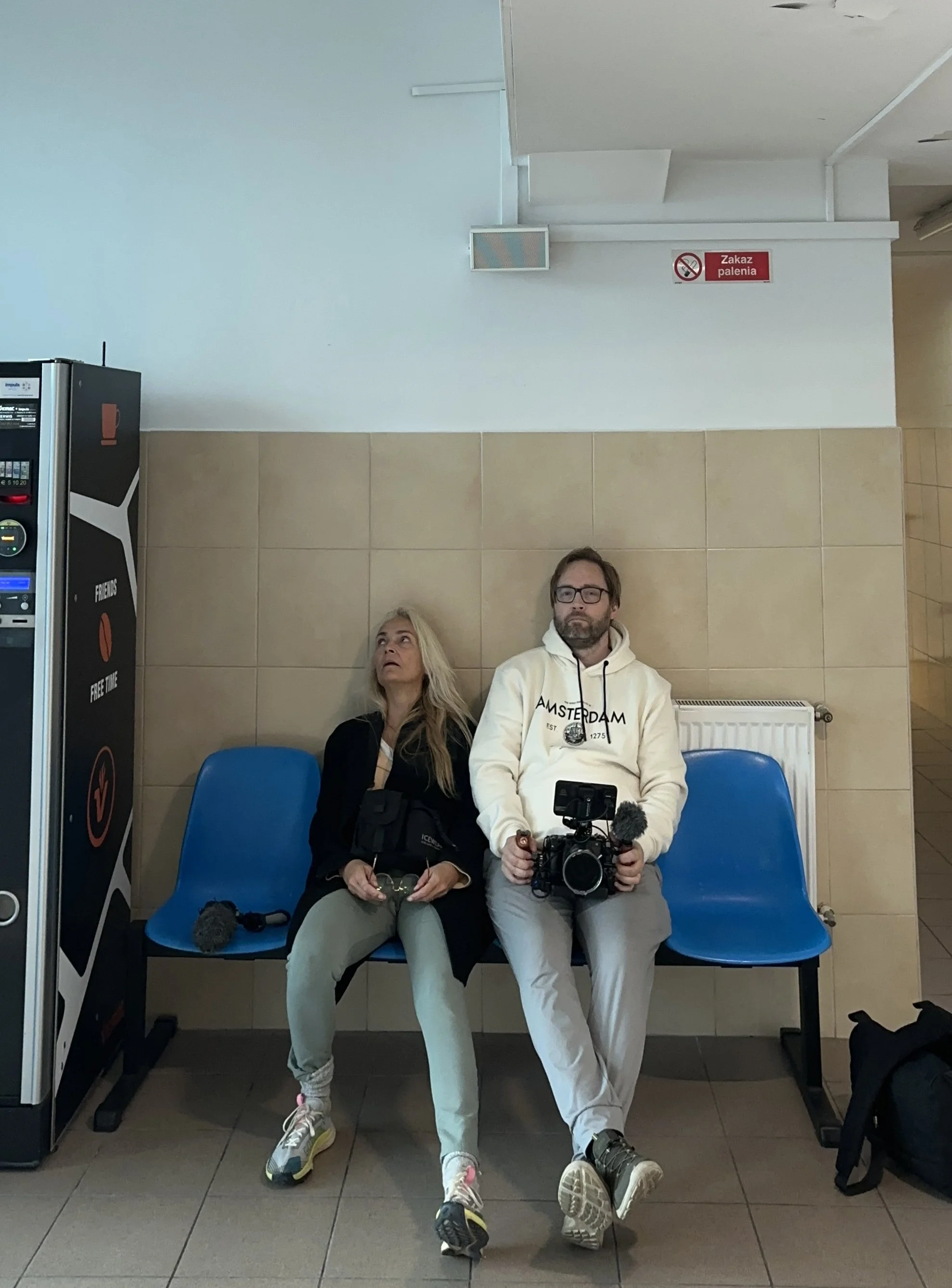 Two people sitting on blue chairs against a beige tile wall in a public space. The woman on the left has long blonde hair, wears glasses, a black jacket, beige pants, and sneakers, and is holding sunglasses. The man on the right has brown hair, a beard, glasses, a white hoodie with 'Amsterdam' written on it, and white pants, and is holding a professional camera. There is a vending machine to the left and a backpack on the floor to the right.