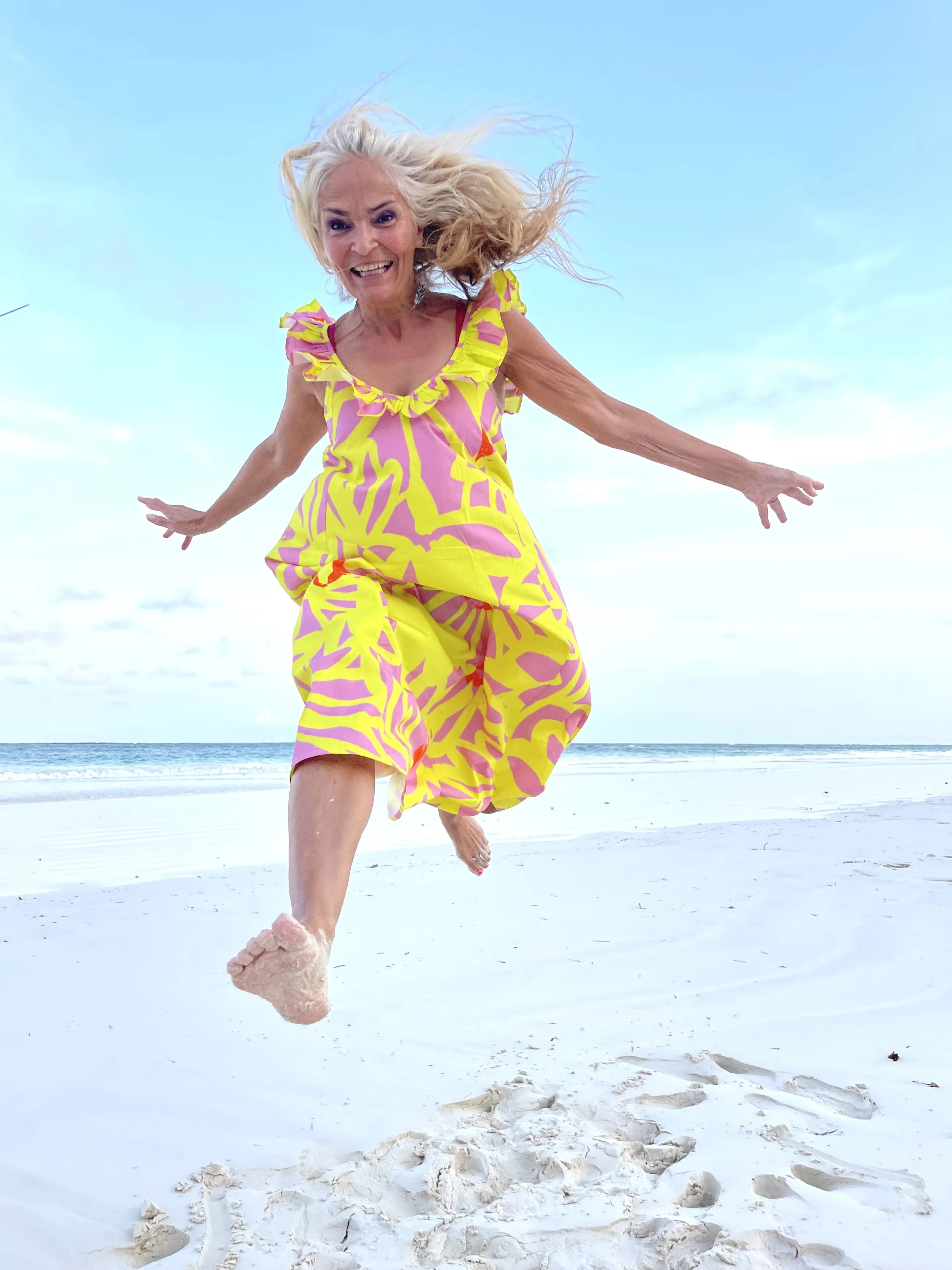 An older woman with gray hair jumping on a sandy beach with the ocean in the background, wearing a colorful pink and yellow patterned summer dress.