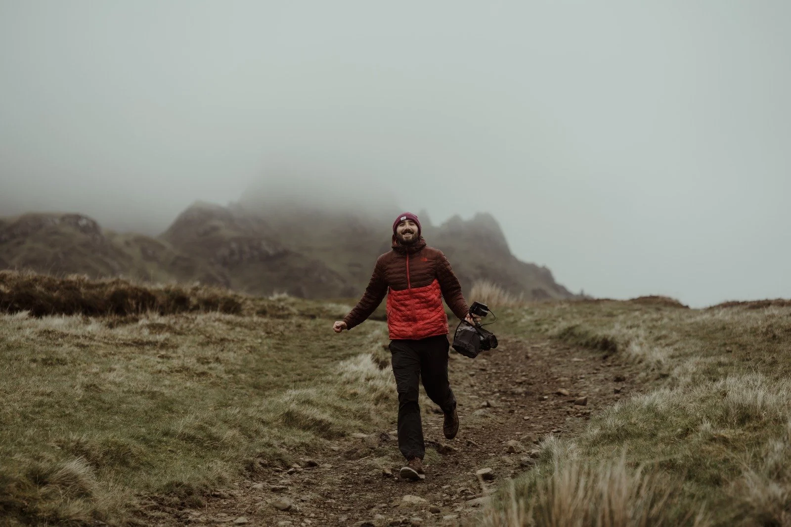 A man in outdoor gear, including a red and brown jacket, black pants, and a beanie, smiling and walking on a dirt trail in a misty, mountainous landscape while carrying a camera.