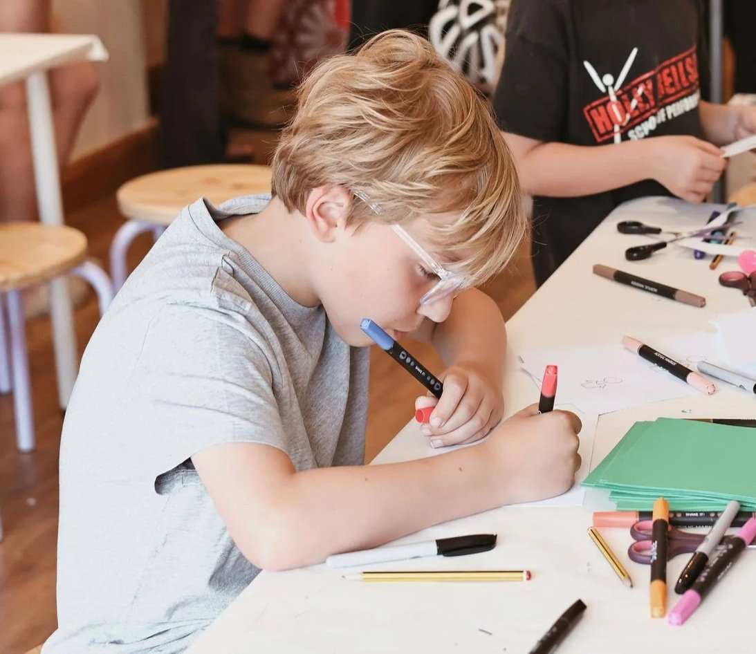 A young boy with blonde hair and glasses drawing with markers at a table covered in various art supplies.