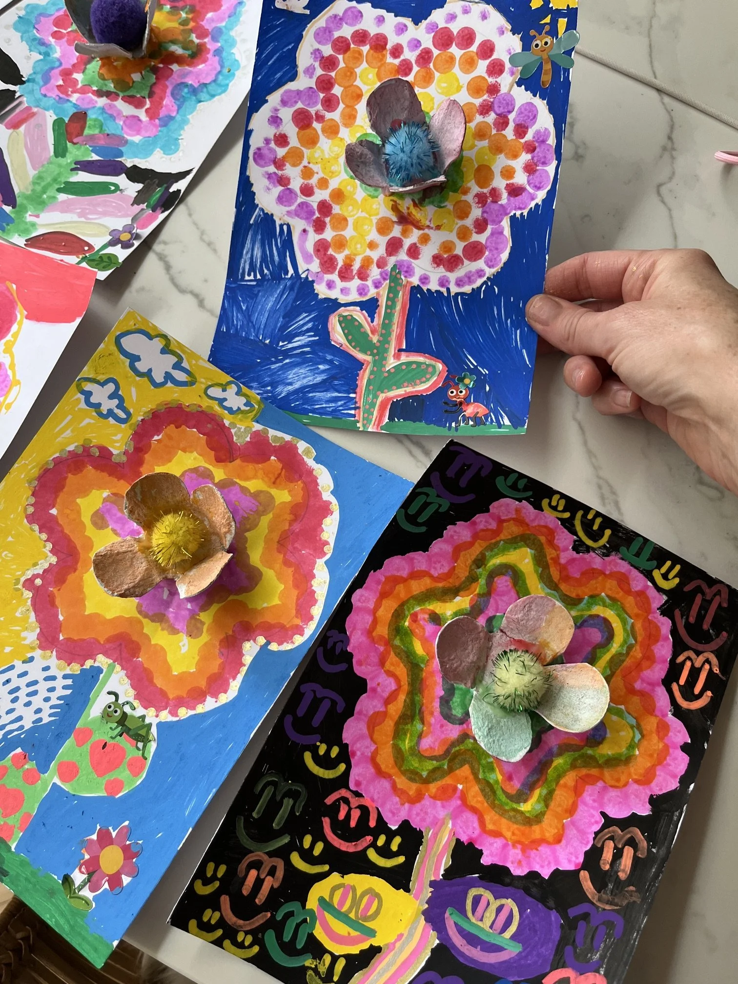 Colorful children's artwork featuring a flower with a blue and green stem, pink, purple, orange, and yellow petals, and a fluffy center. The art is decorated with stickers, 3D flower petals, and vibrant background designs.