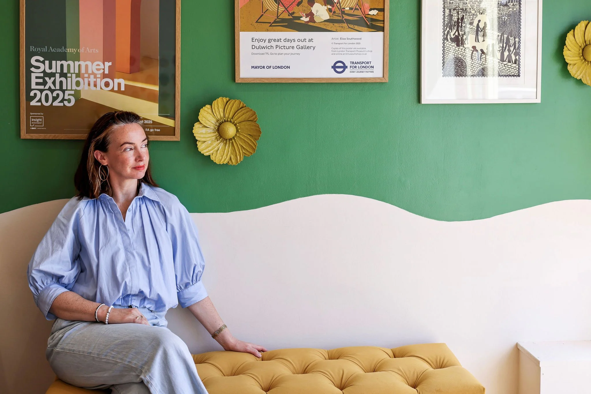 A woman with shoulder-length hair sits on a yellow tufted bench in front of a green wall decorated with framed posters and artwork, wearing a light blue blouse and beige pants.