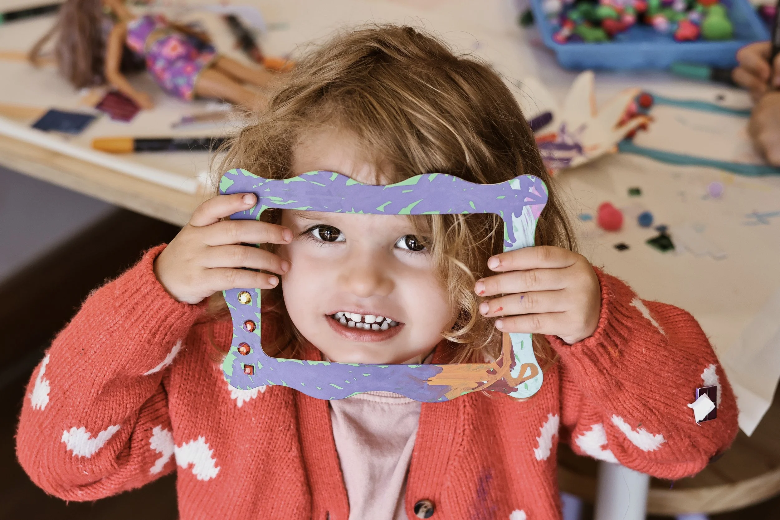 A young girl with curly blond hair and brown eyes holding a colorful, abstract picture frame in front of her face, smiling with teeth visible, wearing a red sweater with white patterns, in a room with art supplies and scattered artwork in the background.