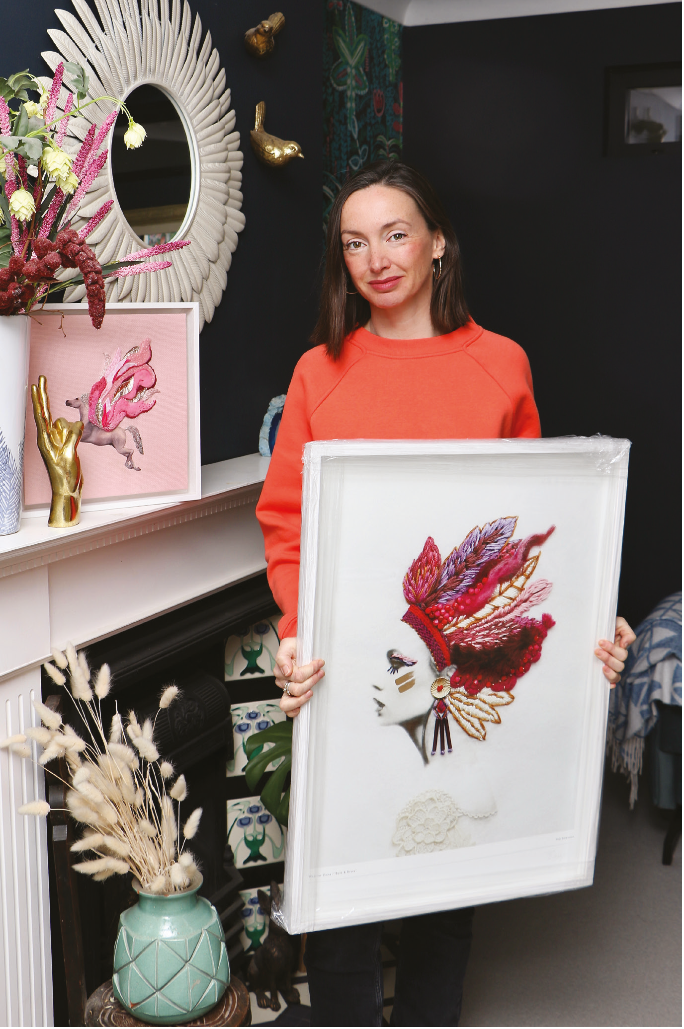 A woman in an orange shirt holding a framed embroidery artwork of a Native American headdress. The background features decorative items, including a large mirror with shell-like frame, golden whale sculptures, colorful art on the wall, and a variety of vases with dried and fresh flowers.