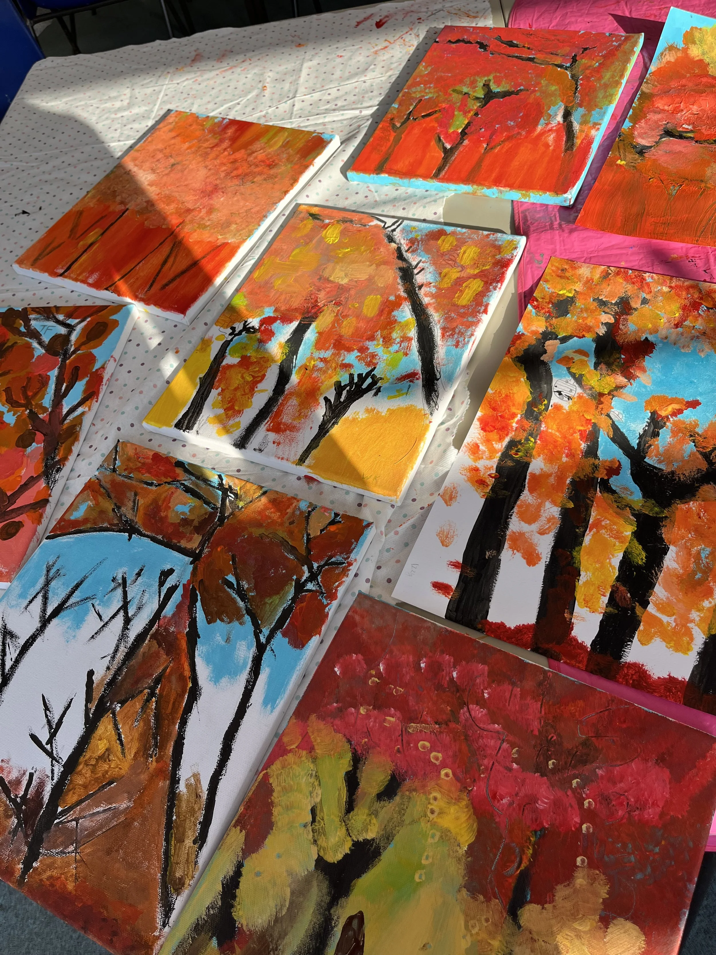 Several colorful abstract landscape paintings on a table, with vibrant autumn trees and backgrounds, painted with thick brushstrokes.