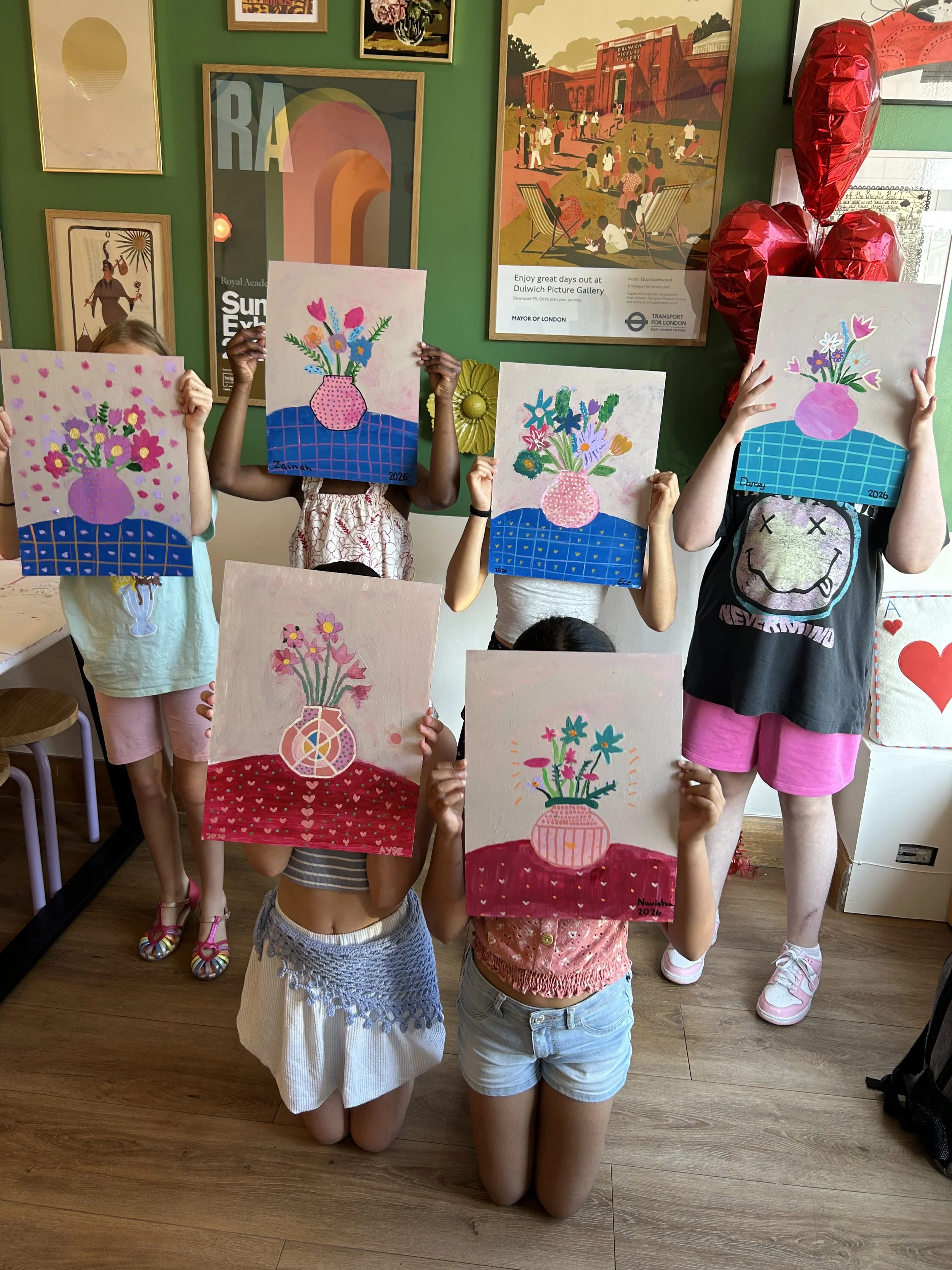 Children holding up colorful paintings of vases with flowers, with a green wall decorated with framed artwork and a large red heart-shaped balloon in the background.
