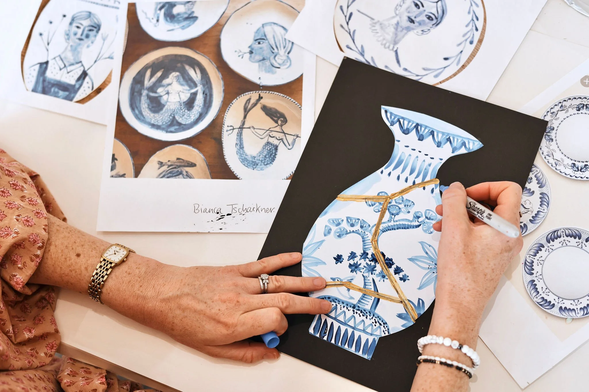 Hands of an artist drawing blue and white porcelain pattern on a ceramic vase illustration with black marker, surrounded by printouts of similar designs.