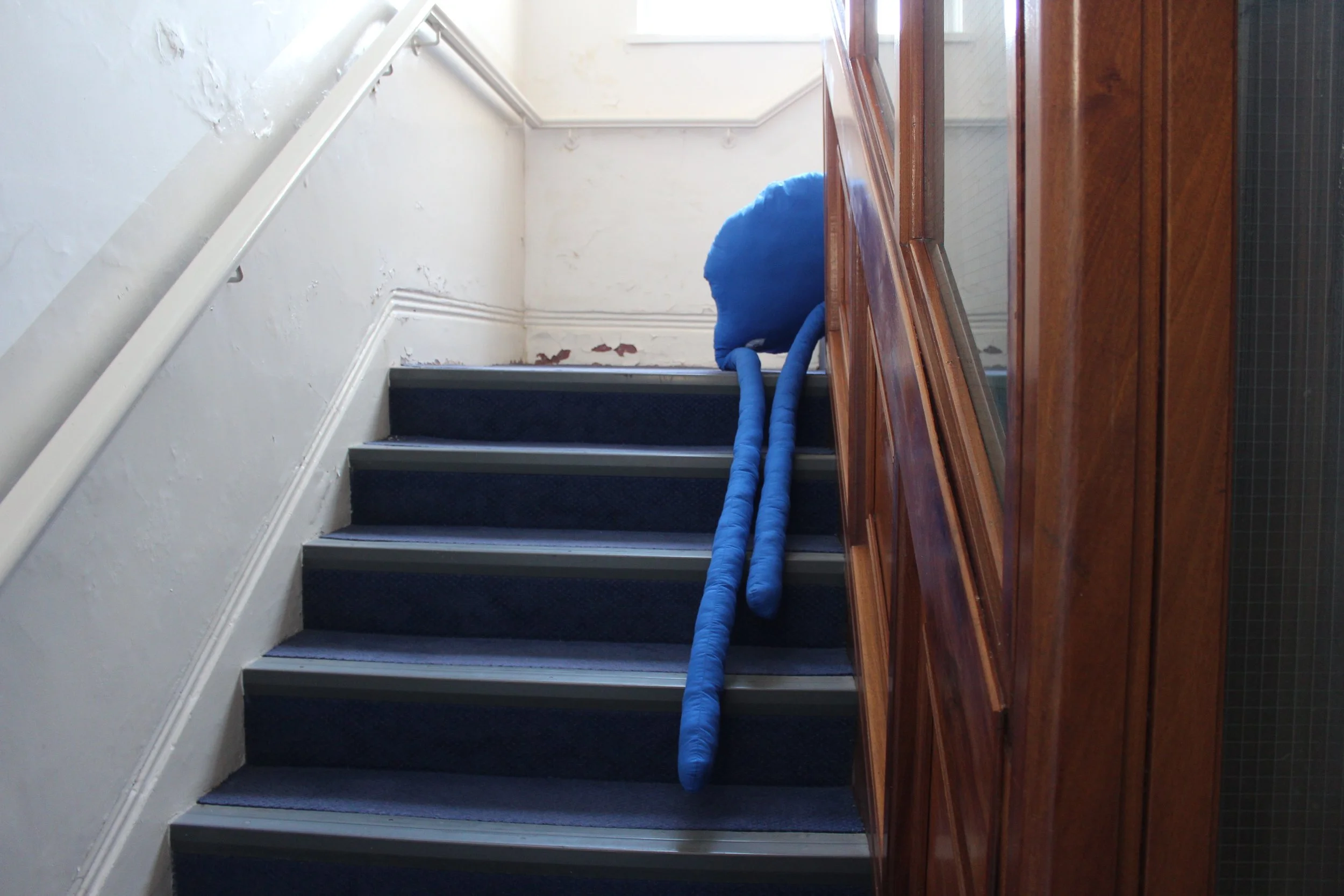 A staircase with dark blue carpeted steps, a white handrail, and a wall with peeling paint. On the right side, a wooden cabinet with glass doors and a dark tiled wall behind it. A large blue flexible duct is draped across the stairs.