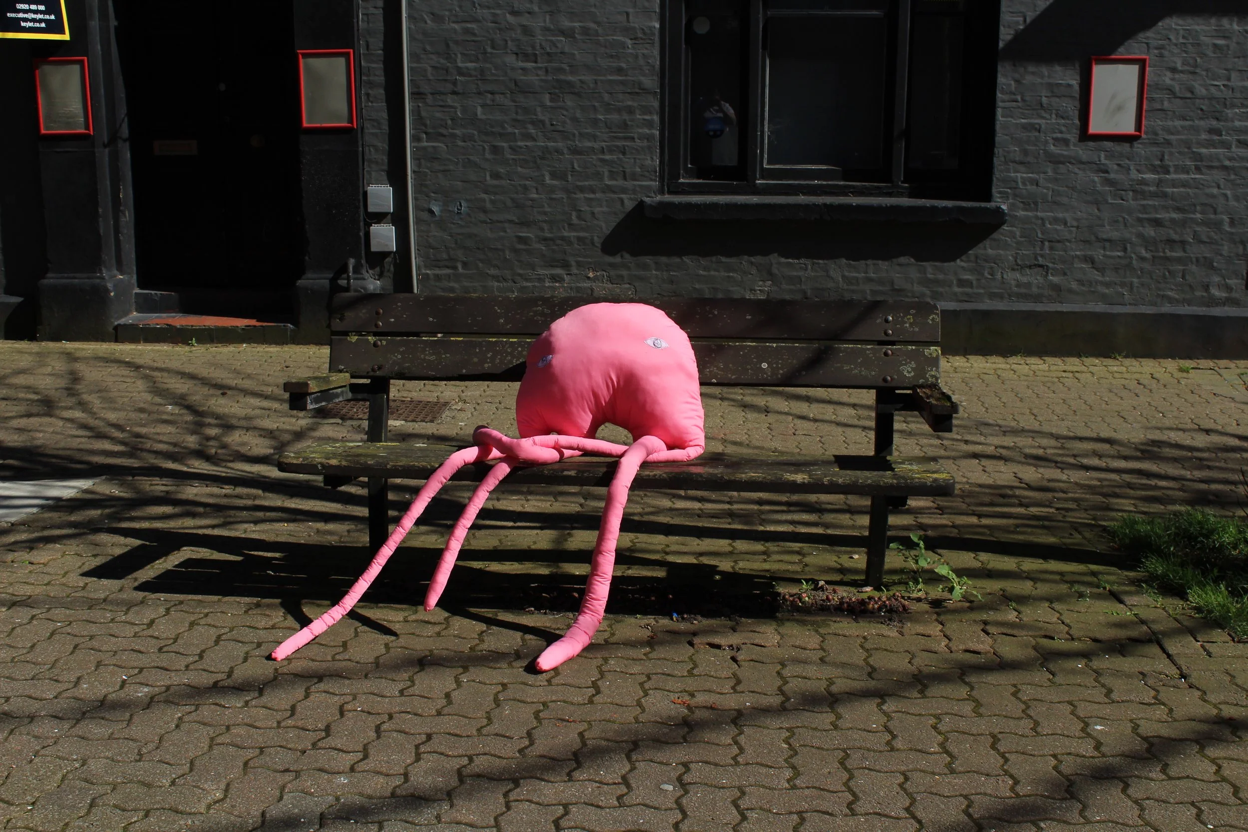 Pink plush octopus toy with long tentacles sitting on a wooden park bench outside a brick building.