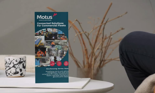 A brochure titled 'Motus' on a white table next to a white mug with black splatter design and a leafless plant with brown twigs, with part of a person's leg in dark clothing visible.