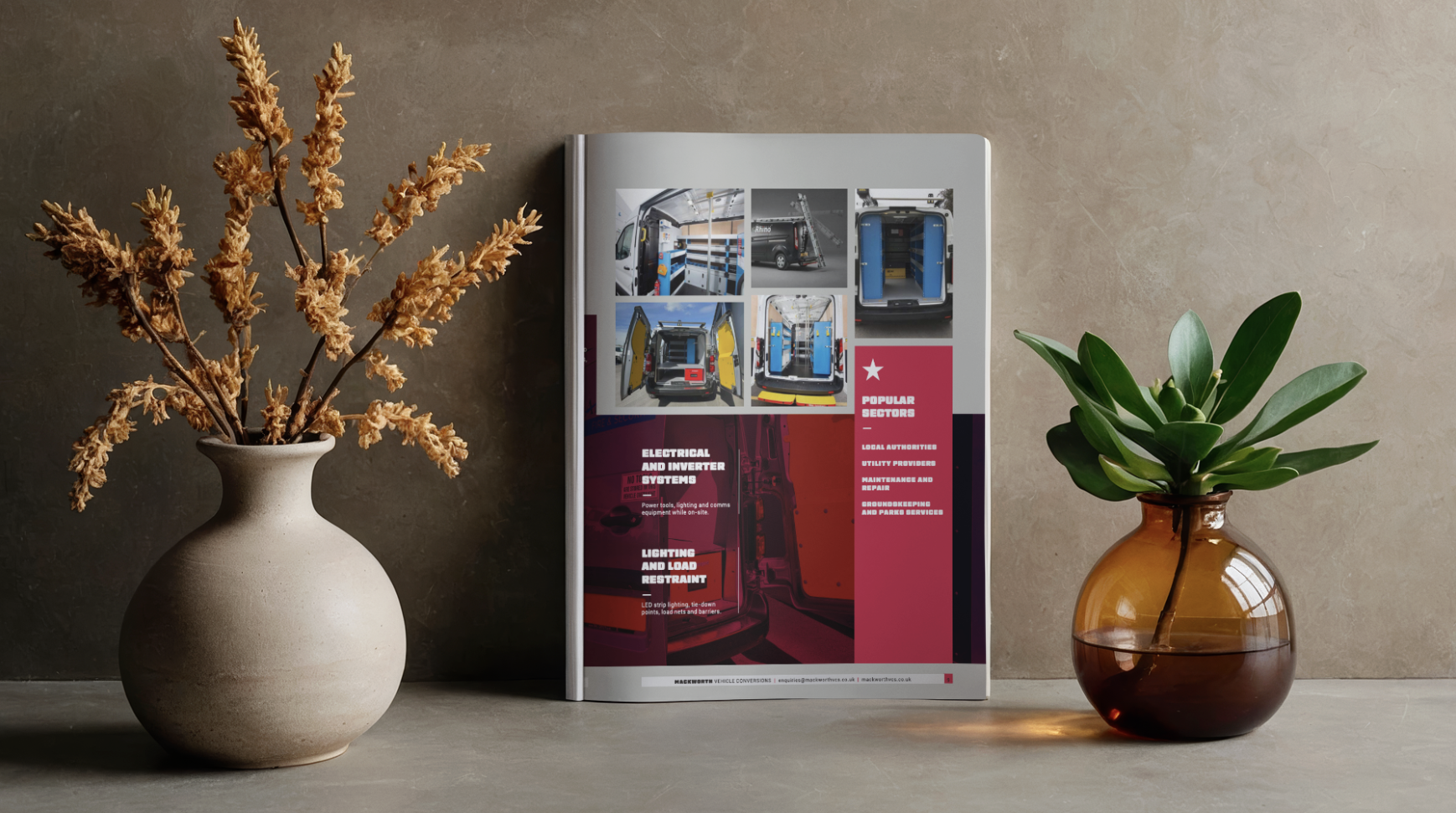 A magazine or brochure standing upright on a table with two vases of flowers on either side. The left vase has dried, brown flowers, while the right vase has green, leafy plants. The magazine features images of vehicle interior modifications and tools, with text related to electrical systems, inverters, and vehicle maintenance.