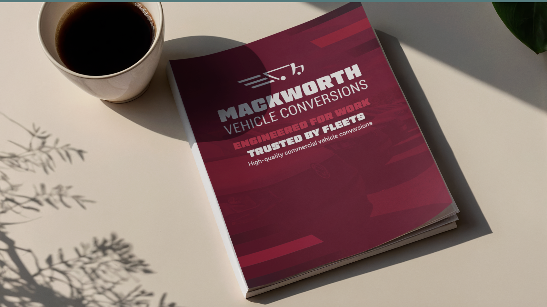 A catalog titled "Mackworth Vehicle Conversions" lying on a white surface next to a cup of coffee. The catalog has a maroon cover with white and red text, and shadows of tree branches are cast on the surface.