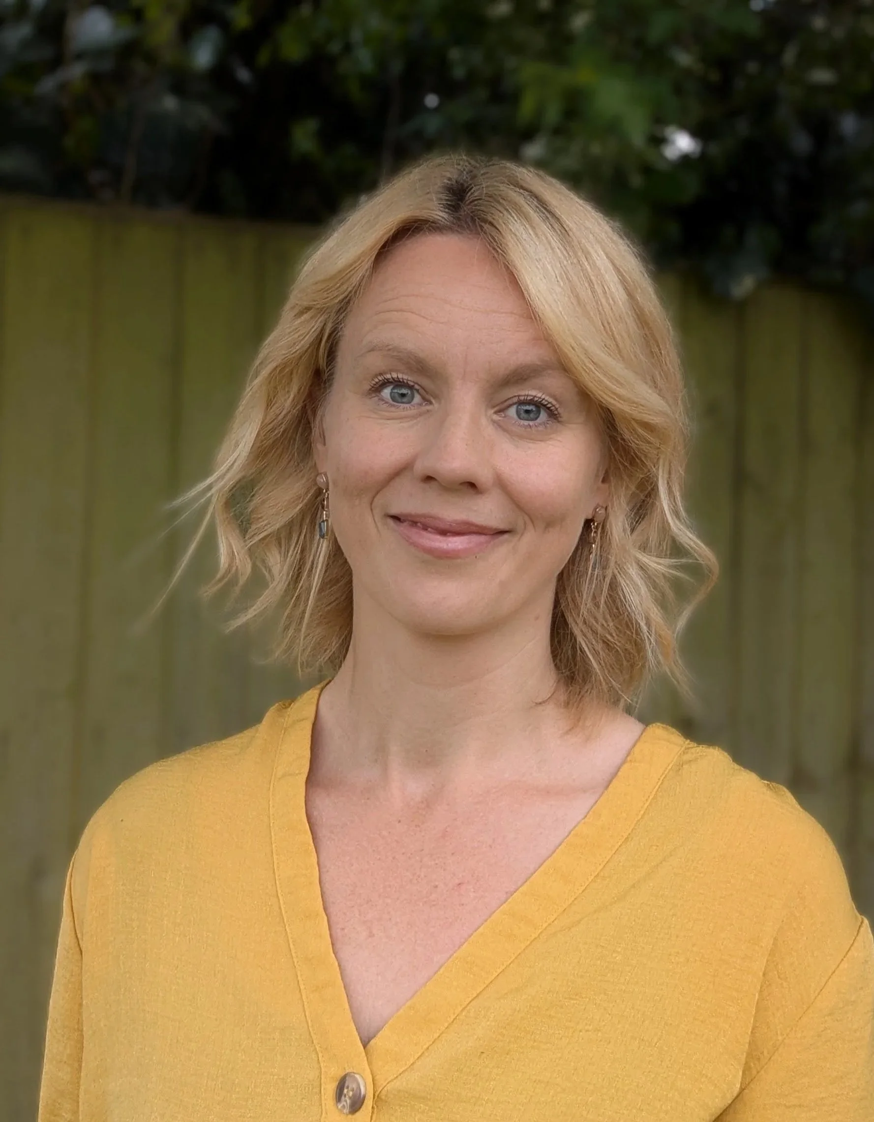 A woman with blonde hair and blue eyes, wearing a yellow V-neck sweater, standing outdoors with a wooden fence and green foliage in the background.