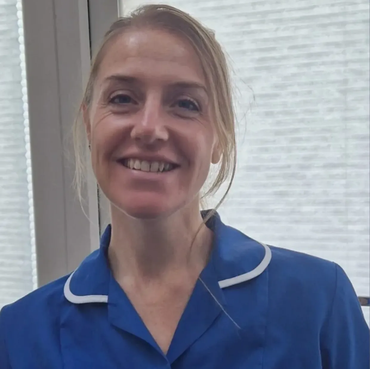 A woman with blonde hair and a bright smile wearing a blue shirt, standing in front of a window with blinds.