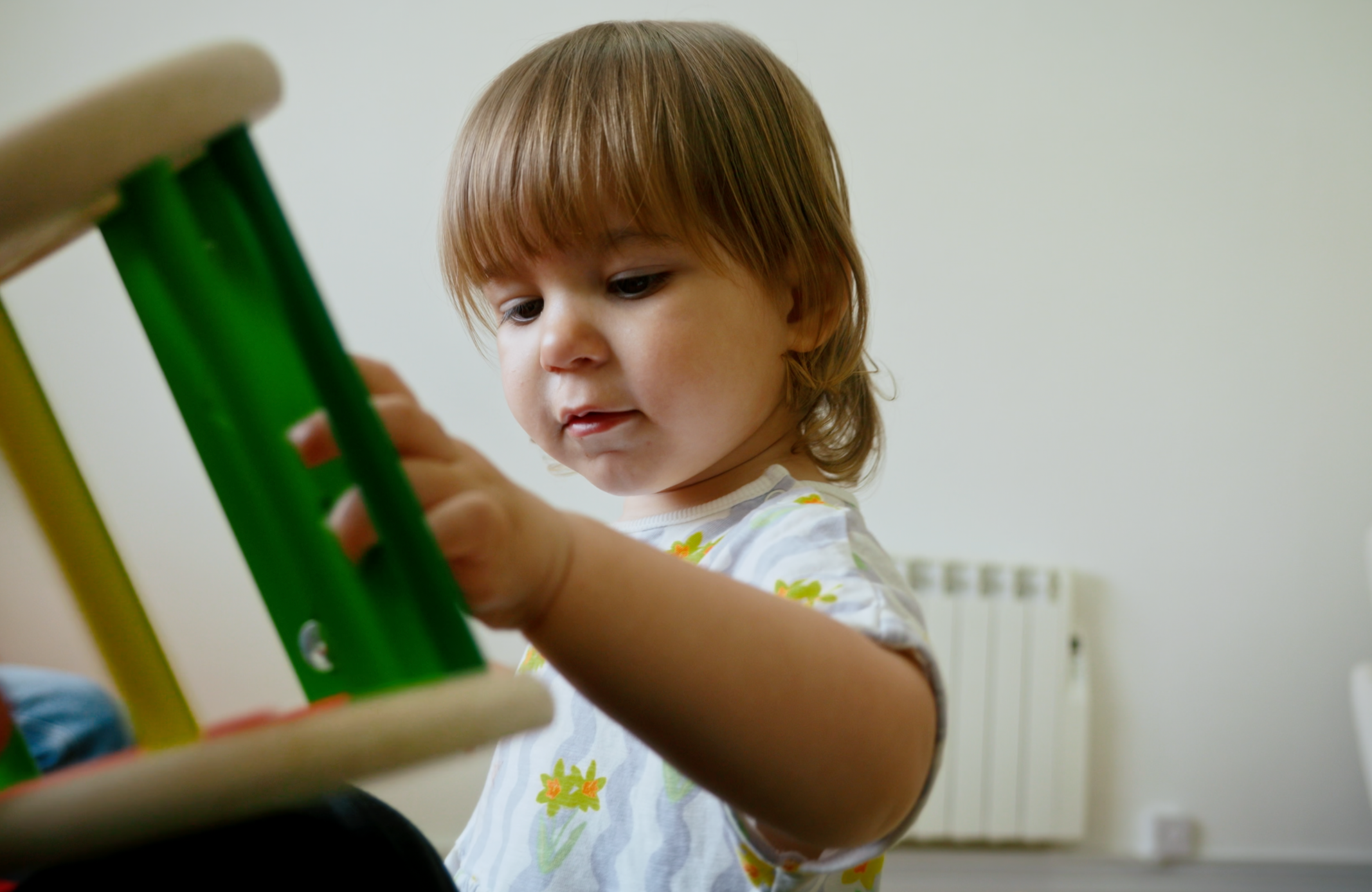 A young child with light brown hair wearing a white shirt with yellow and green floral pattern, examining or holding a small object, with a plain white wall and radiator in the background.